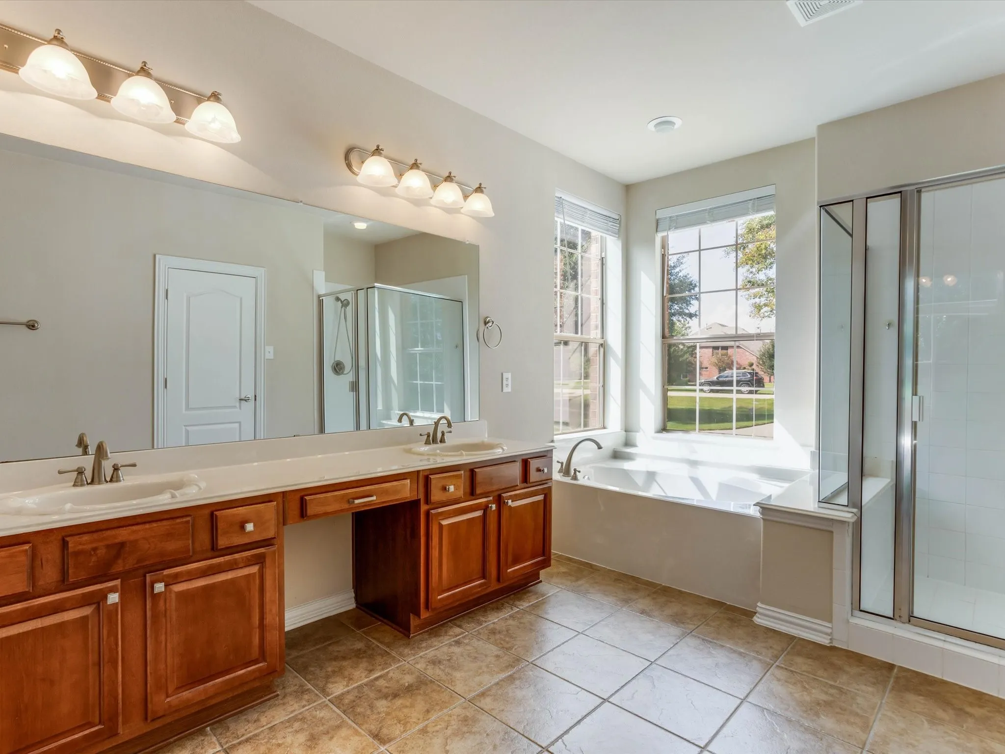Dual-sink vanity with wood-finish cabinetry, solid-surface countertops, soaking tub, glass-enclosed shower, and tile flooring