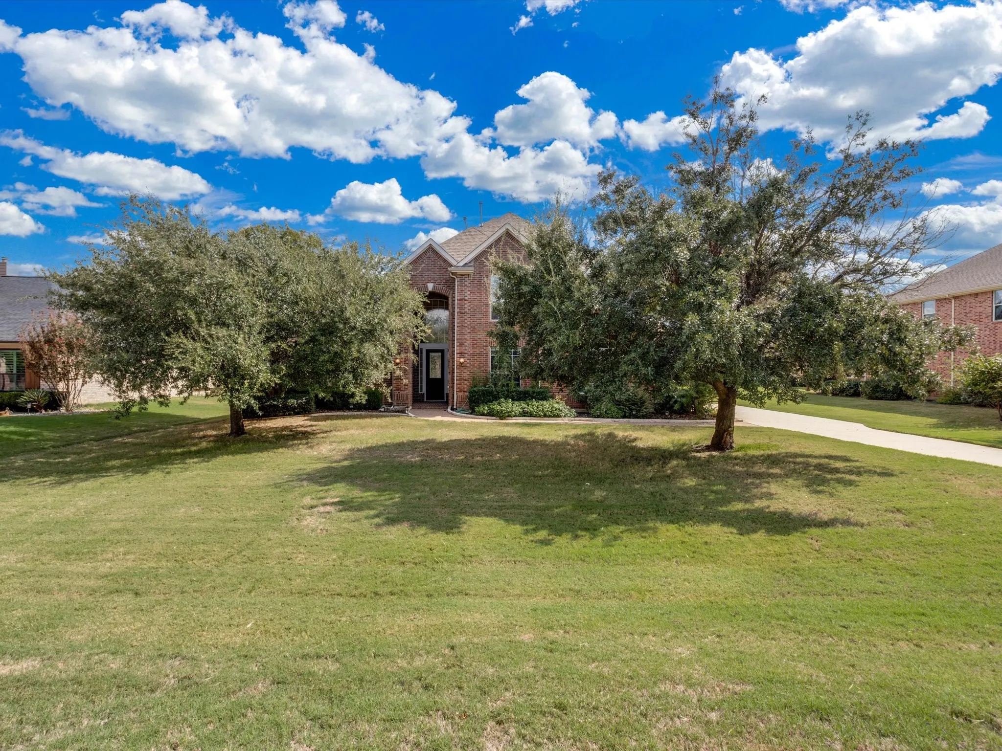 Brick exterior residence featuring a steeply pitched roof, arched entryway, concrete driveway, and mature landscaping