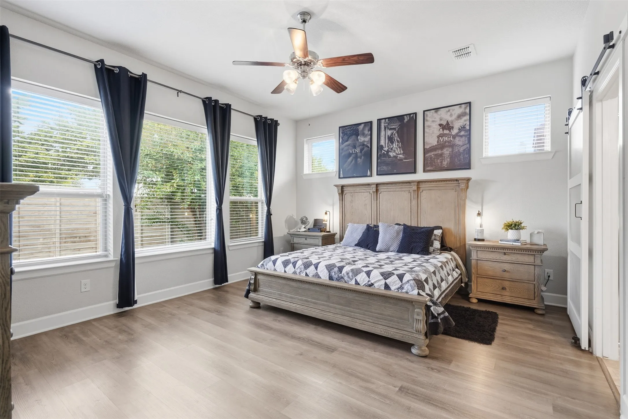 Spacious room featuring wood-finish flooring, high ceilings with a central multi-blade light fixture, expansive windows, and a white sliding barn-style door to   the bathroom.