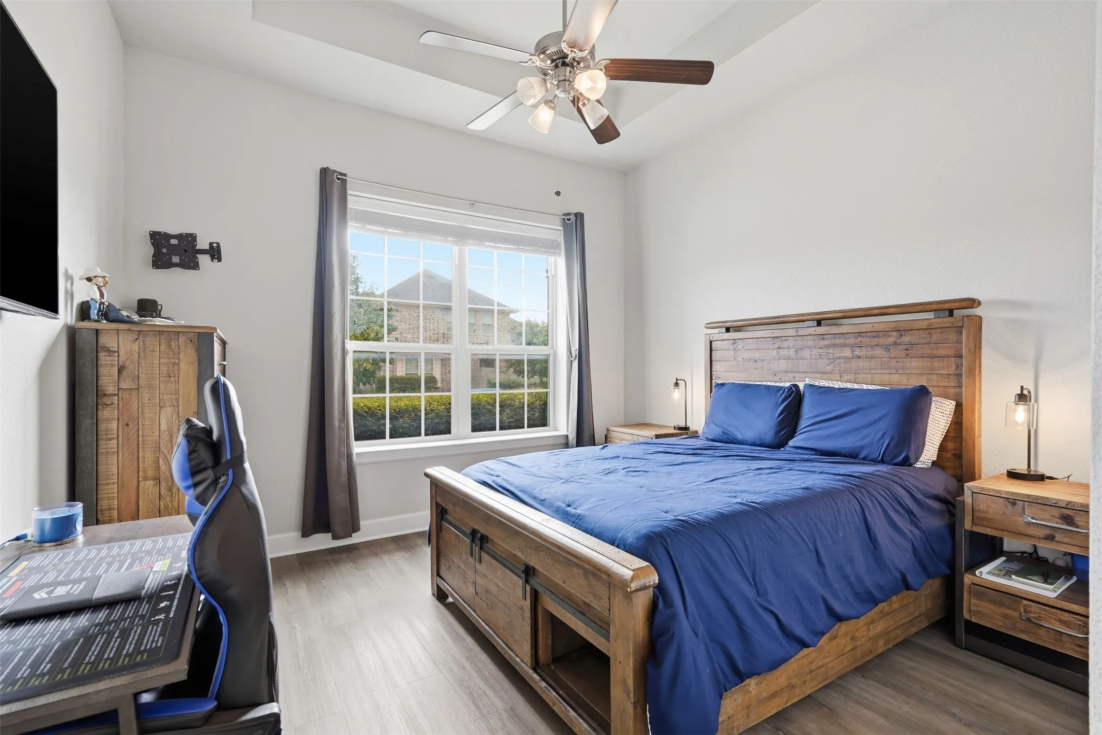 Room featuring a tray ceiling, multi-blade ceiling fan with integrated light fixture, wood-finish flooring, and a large window curtains in front of home.