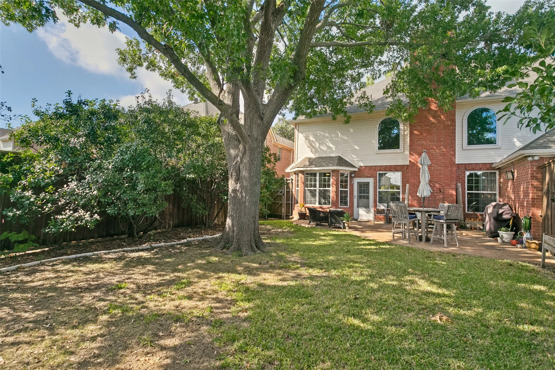 Brick Colonial home with expansive rear grounds featuring mature shade trees, manicured lawn, and covered patio with outdoor seating area