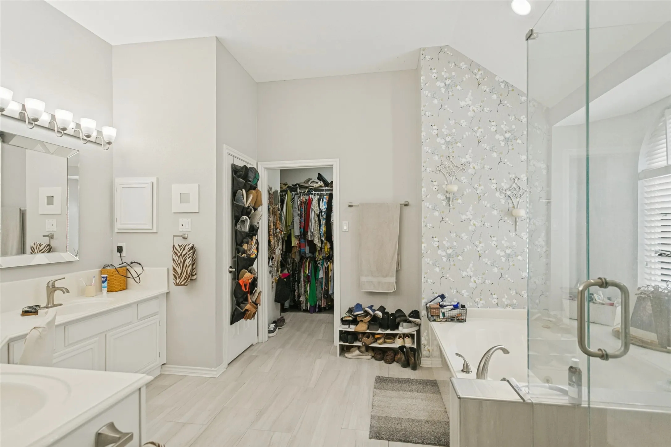 Bathroom featuring a dual-sink vanity with white cabinetry, a glass-enclosed shower, a deep soaking tub, and ceramic tile flooring