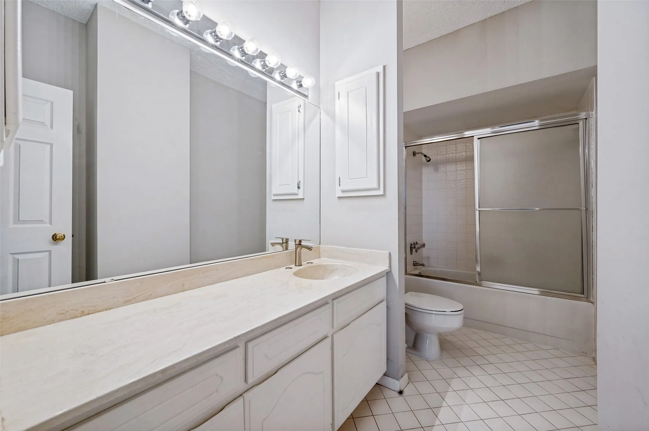 Bathroom vanity with a stone countertop, integrated basin, and full-width wall mirror, accompanied by a shower-tub combination featuring a sliding glass enclosure and tiled surround