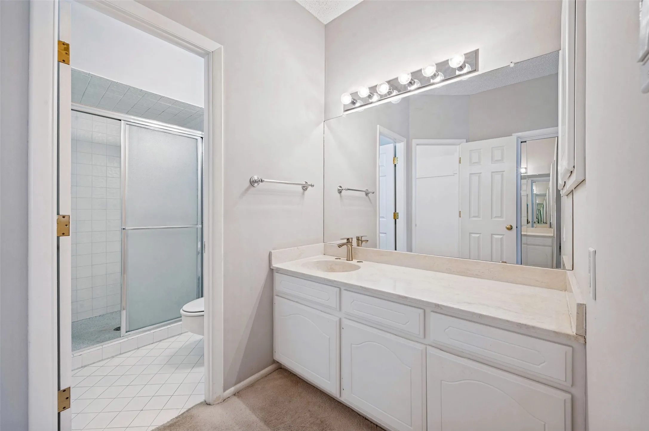 Bathroom featuring a white vanity with a neutral stone countertop, an expansive wall-mounted mirror, multi-bulb vanity lighting, and a glass-enclosed shower stall with tile flooring