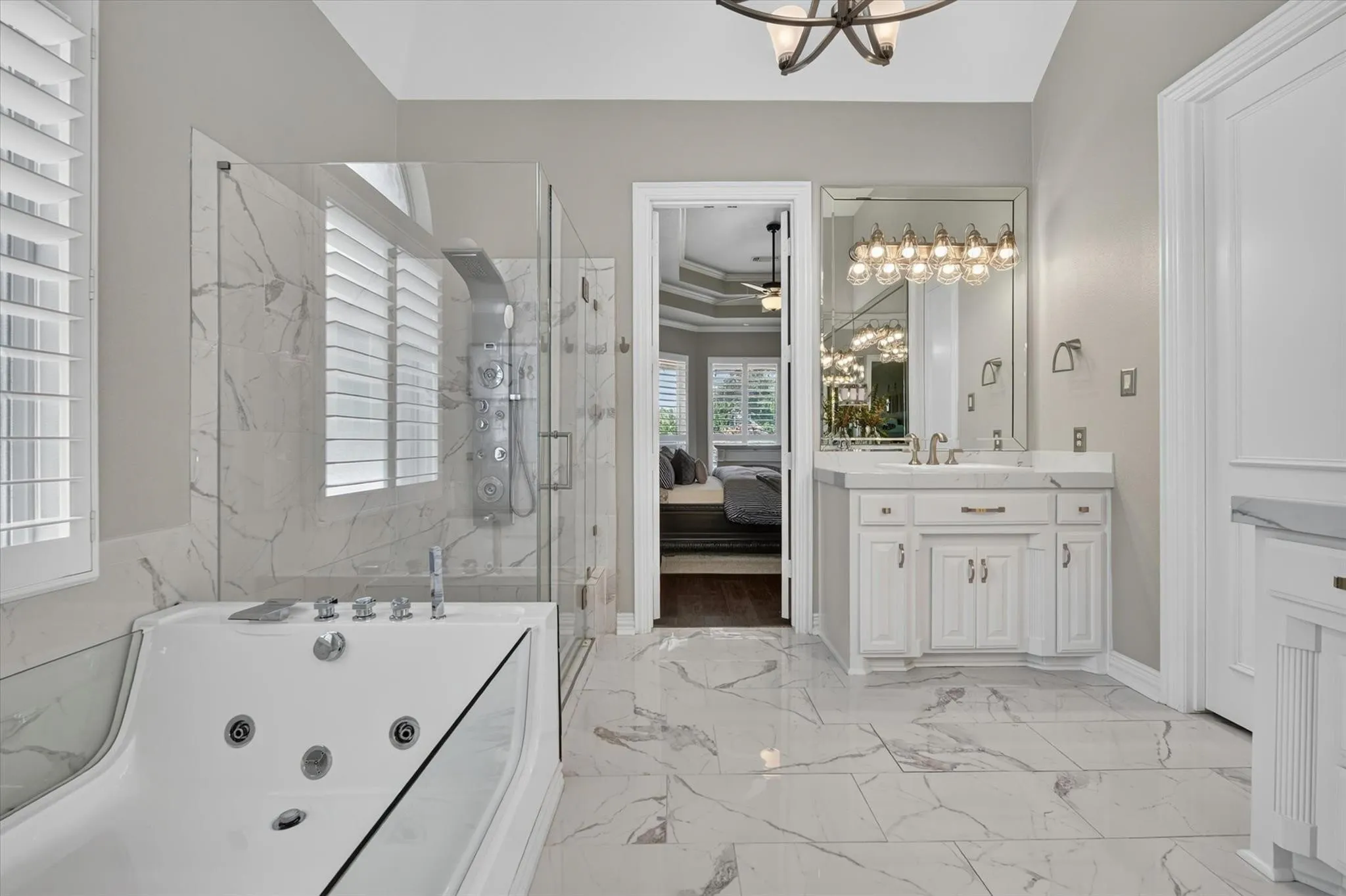 Bathroom featuring large-format tile flooring, jetted soaking tub, glass-enclosed shower with body spray system, white cabinetry with stone countertops, and a wall-to-wall vanity mirror with integrated lighting