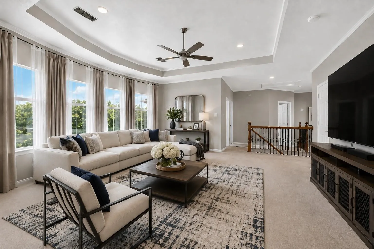 Tray ceiling featuring a central multi-blade ceiling fan, expansive windows, recessed lighting, and a staircase with stained wood handrails and iron balusters