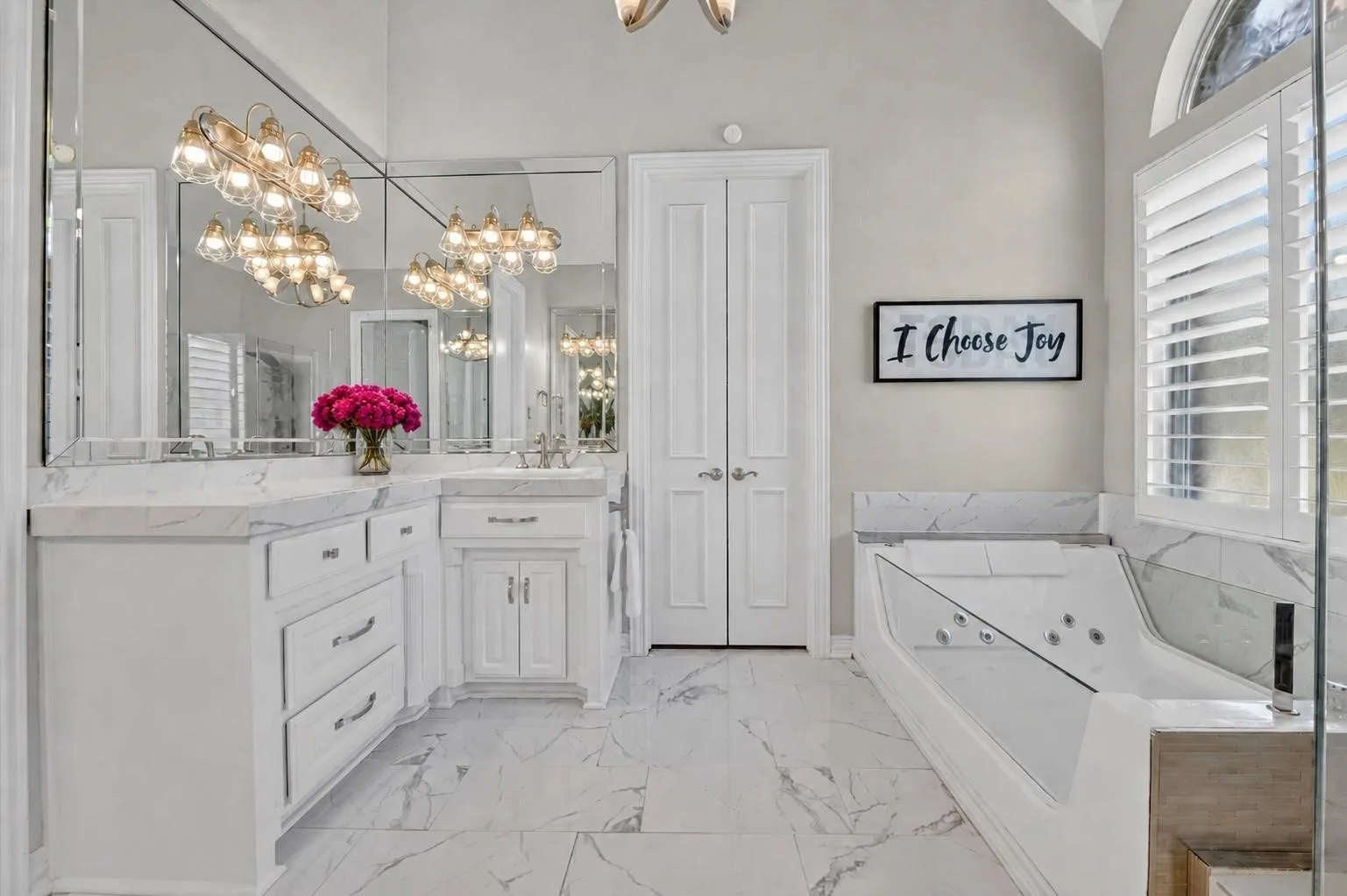 White cabinetry with light-toned countertops, stone-patterned tile flooring, full-width vanity mirror, built-in jetted tub with stone-patterned surround, and arched window with louvered shutters