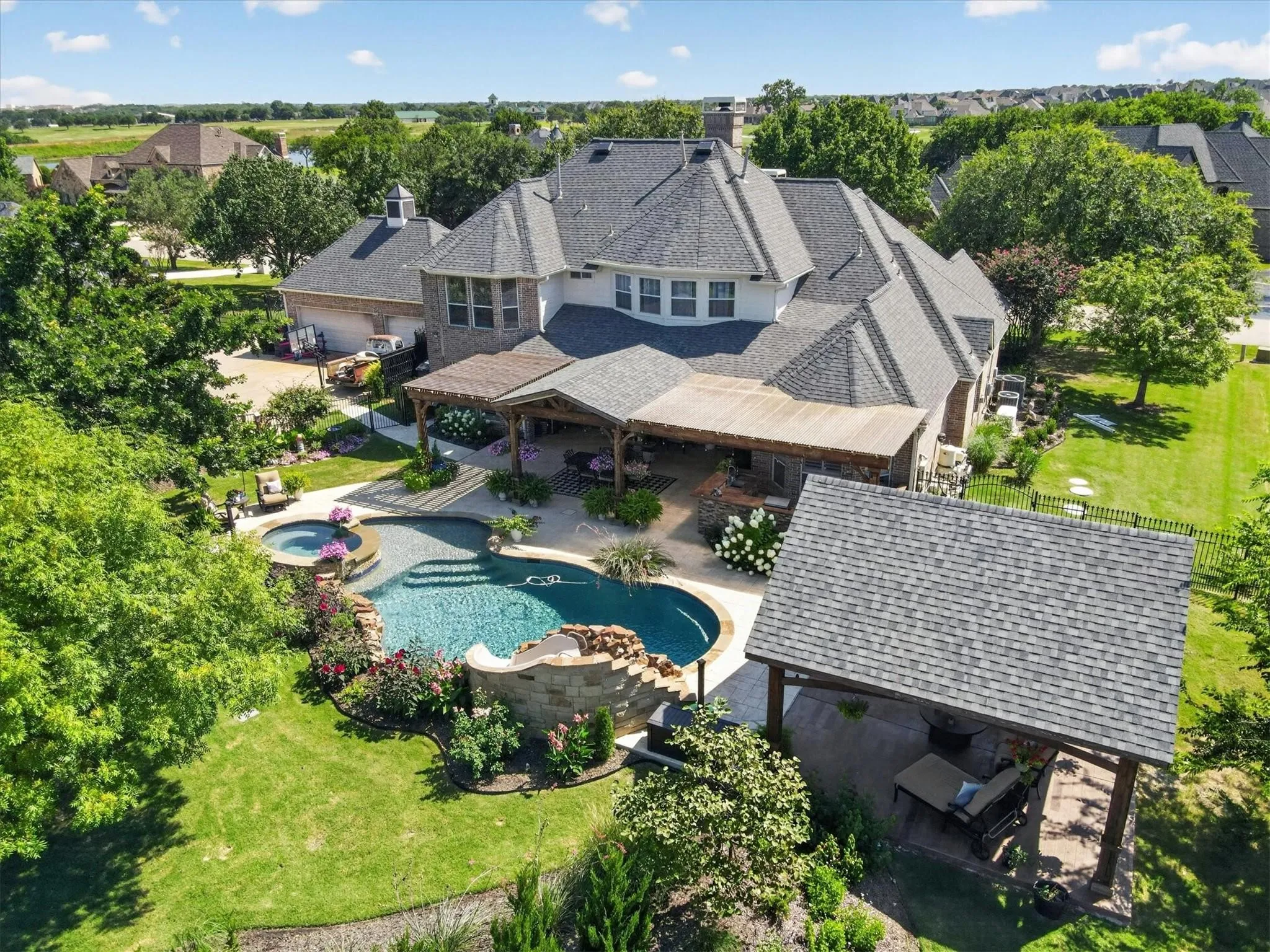 Rear exterior featuring a multi-level stone pool with integrated spa and rock water feature, expansive covered patio with built-in kitchen, detached timber-frame pavilion, and mature landscaping
