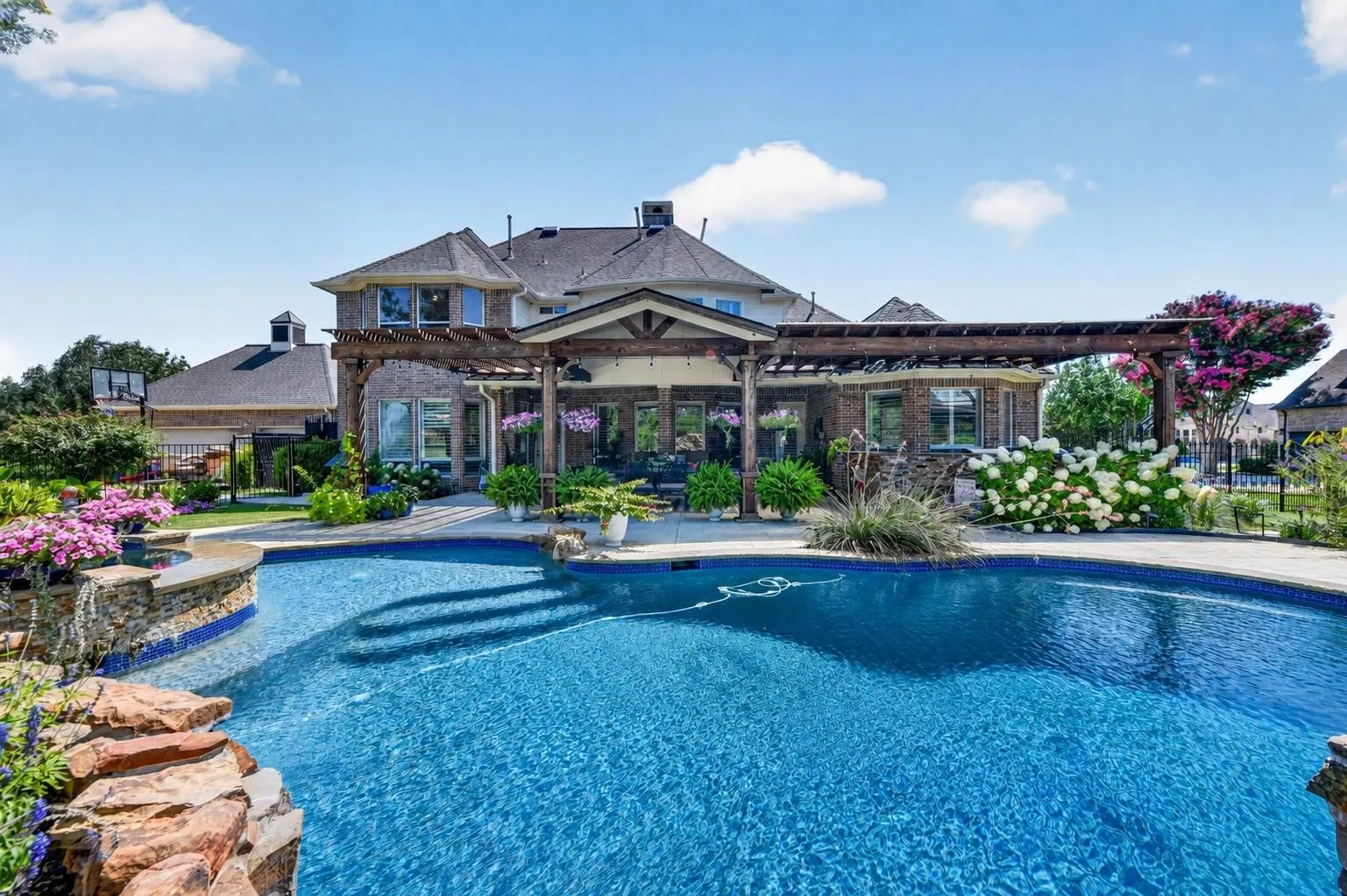 Brick exterior featuring a multi-level roofline, extensive covered pergola with timber framing, custom-shaped swimming pool, and stone masonry waterfall element