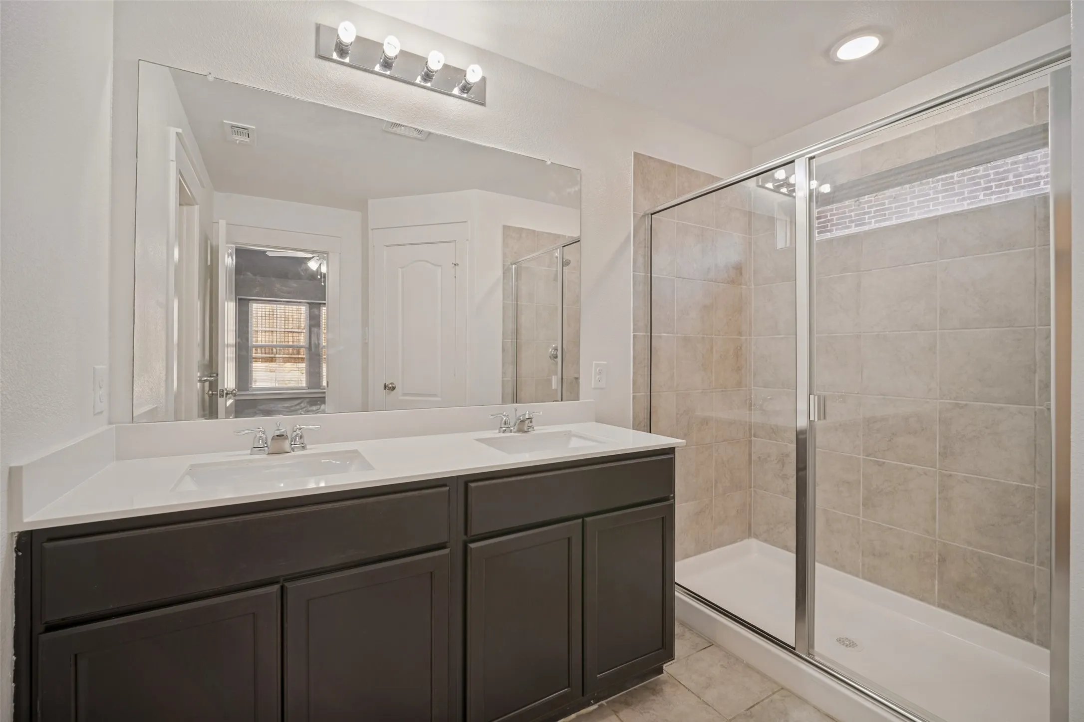 Dual-sink vanity with a solid-surface countertop and charcoal-finish cabinetry, featuring a frameless-style mirror, chrome hardware, and a walk-in shower with tiled surround