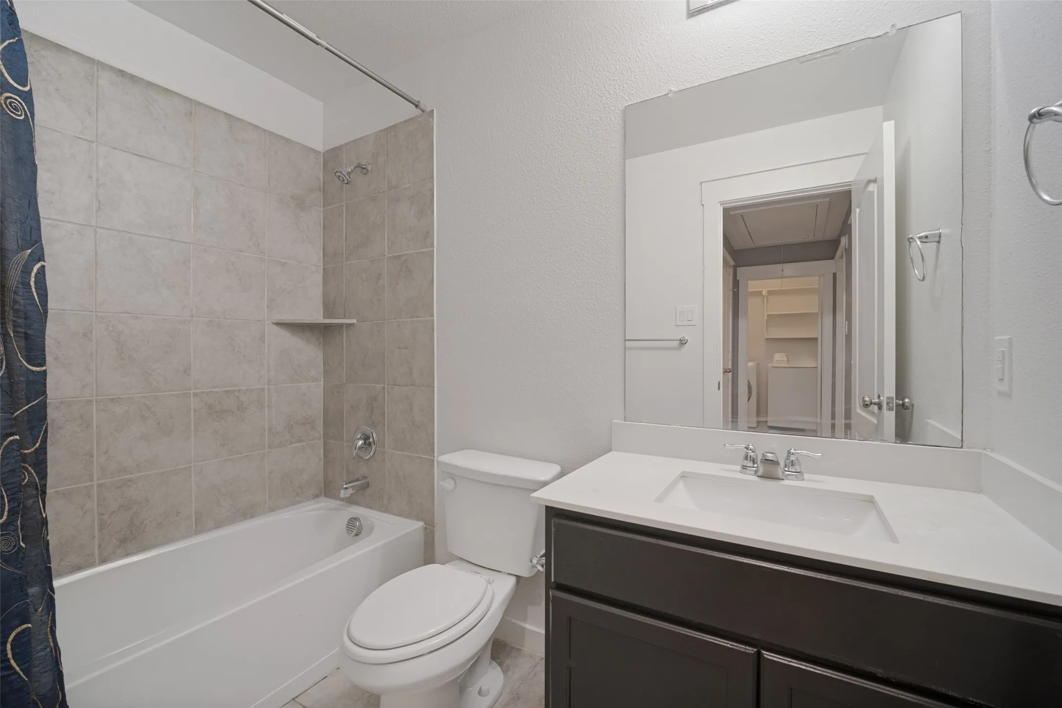 Bathroom featuring a vanity with a white countertop and rectangular undermount sink, dark-toned cabinetry, a full-width wall-mounted mirror, and a tub with large-format stone-style wall tile
