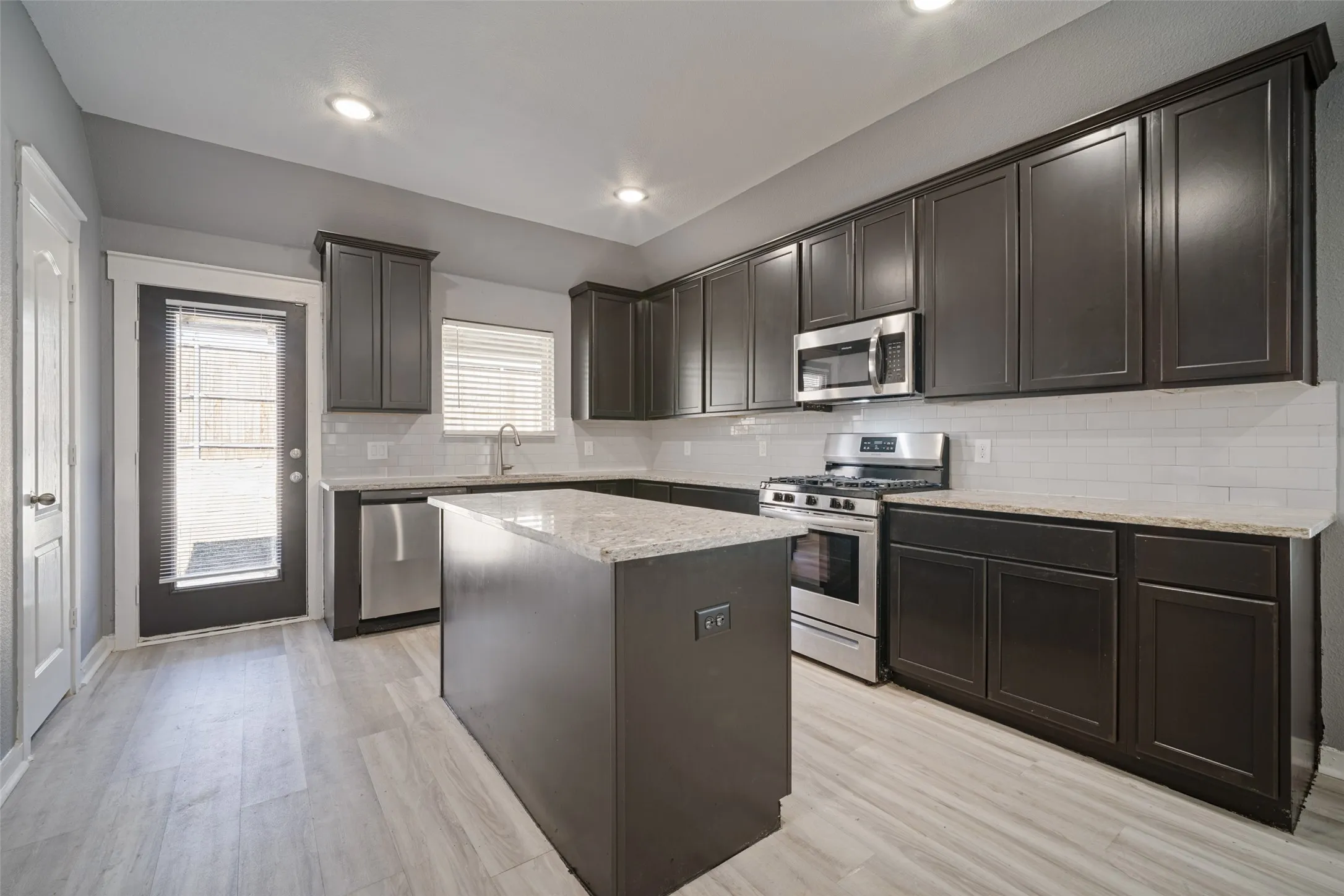 Kitchen featuring dark-finish cabinetry, light-toned stone-style countertops, white subway tile backsplash, stainless steel appliances, and wood-finish flooring