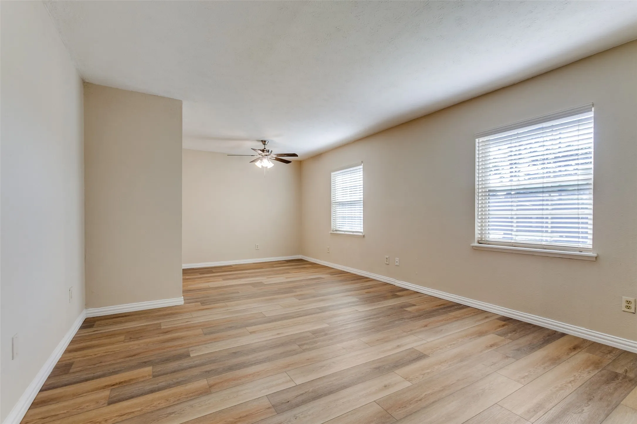 Wood-finish flooring, neutral wall tones, two windows with horizontal blinds, and a multi-blade ceiling fan with integrated lighting