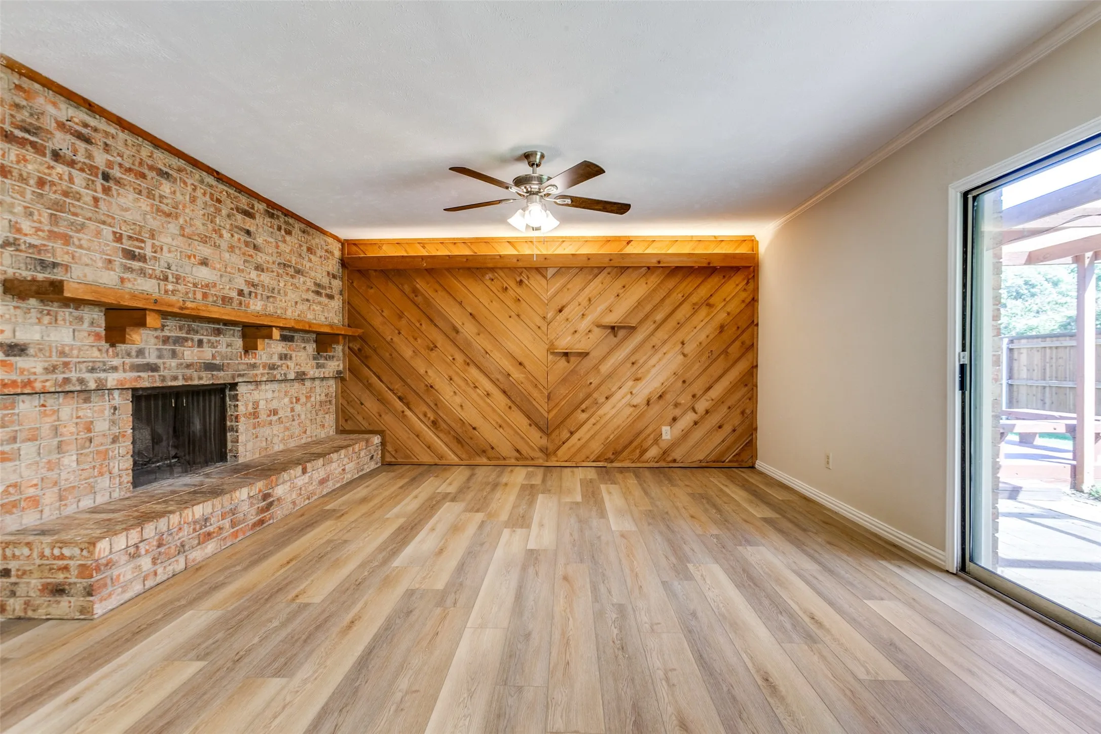 Living area featuring wood-finish flooring, a floor-to-ceiling brick fireplace with a custom wood mantel, diagonal wood-paneled accent wall, and a sliding glass door