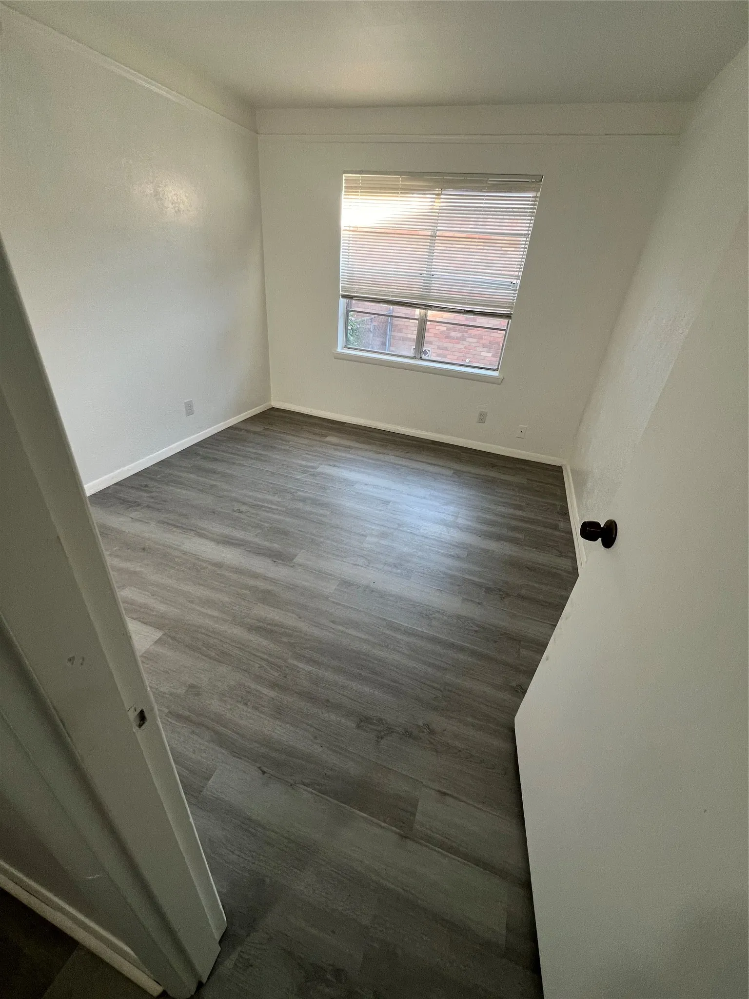 Neutral interior room featuring wood-finish flooring, a single-pane window with horizontal blinds, and a standard swinging door with a matte black knob