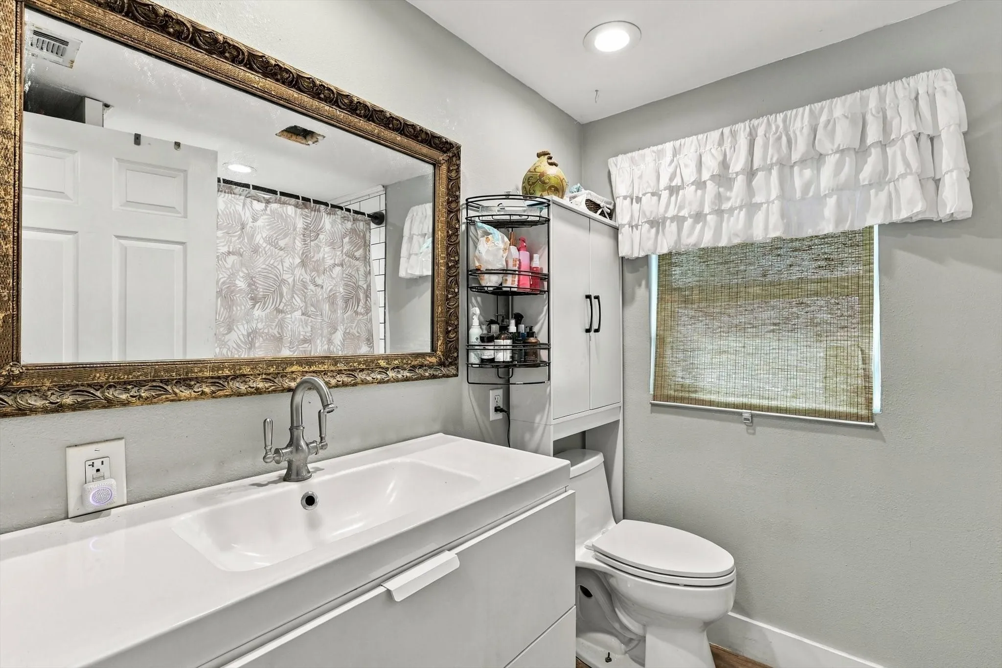 Modern vanity with integrated basin and polished metal faucet, large wall-mounted ornate gold-tone framed mirror, recessed lighting, and built-in vertical storage cabinet