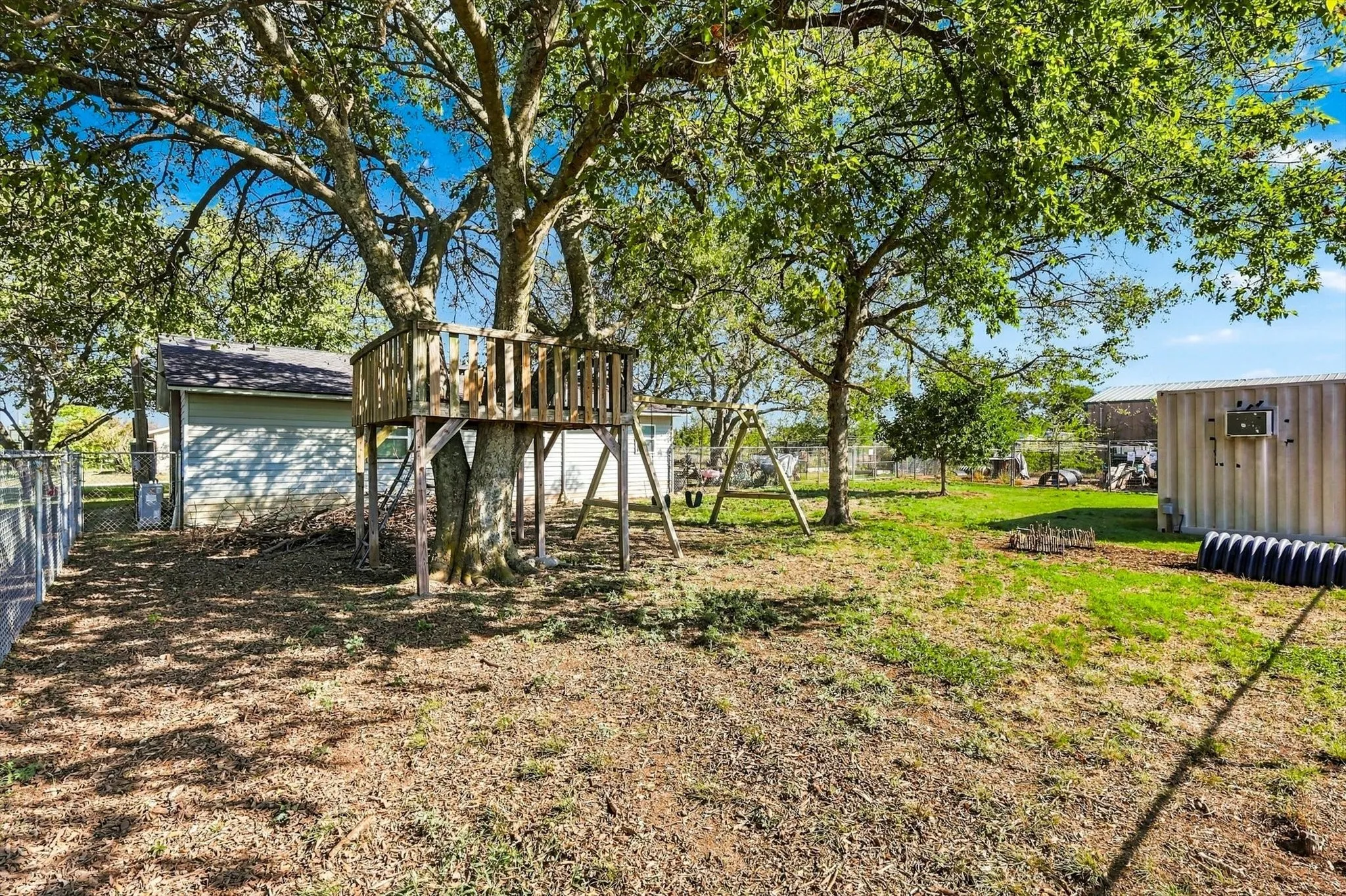 Backyard area featuring mature hardwood trees, a wood-frame tree house and playground, and chain link perimeter fencing