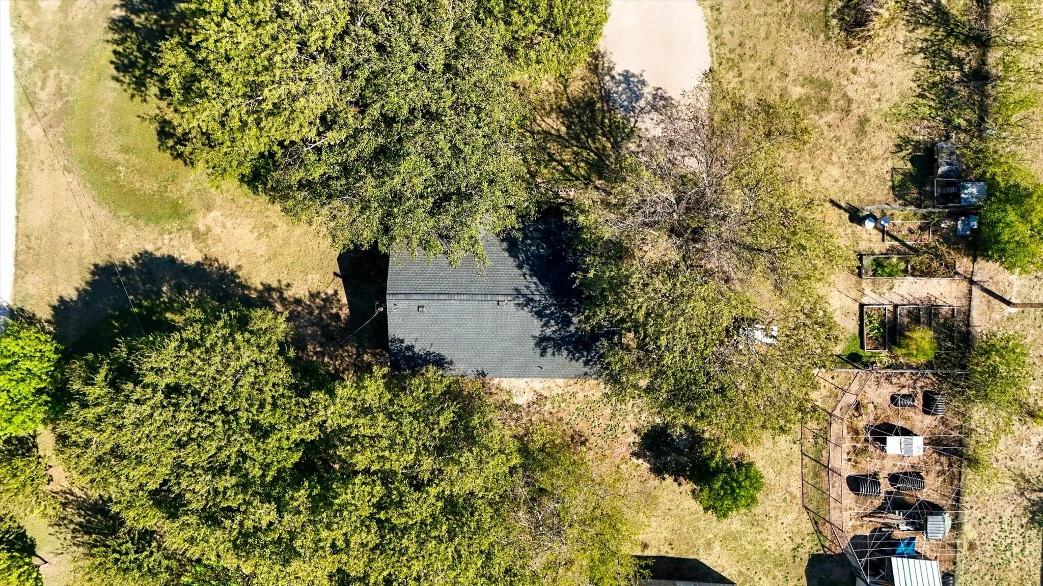 Aerial perspective featuring a shingled roofline, mature trees, open grounds, and a fenced garden area