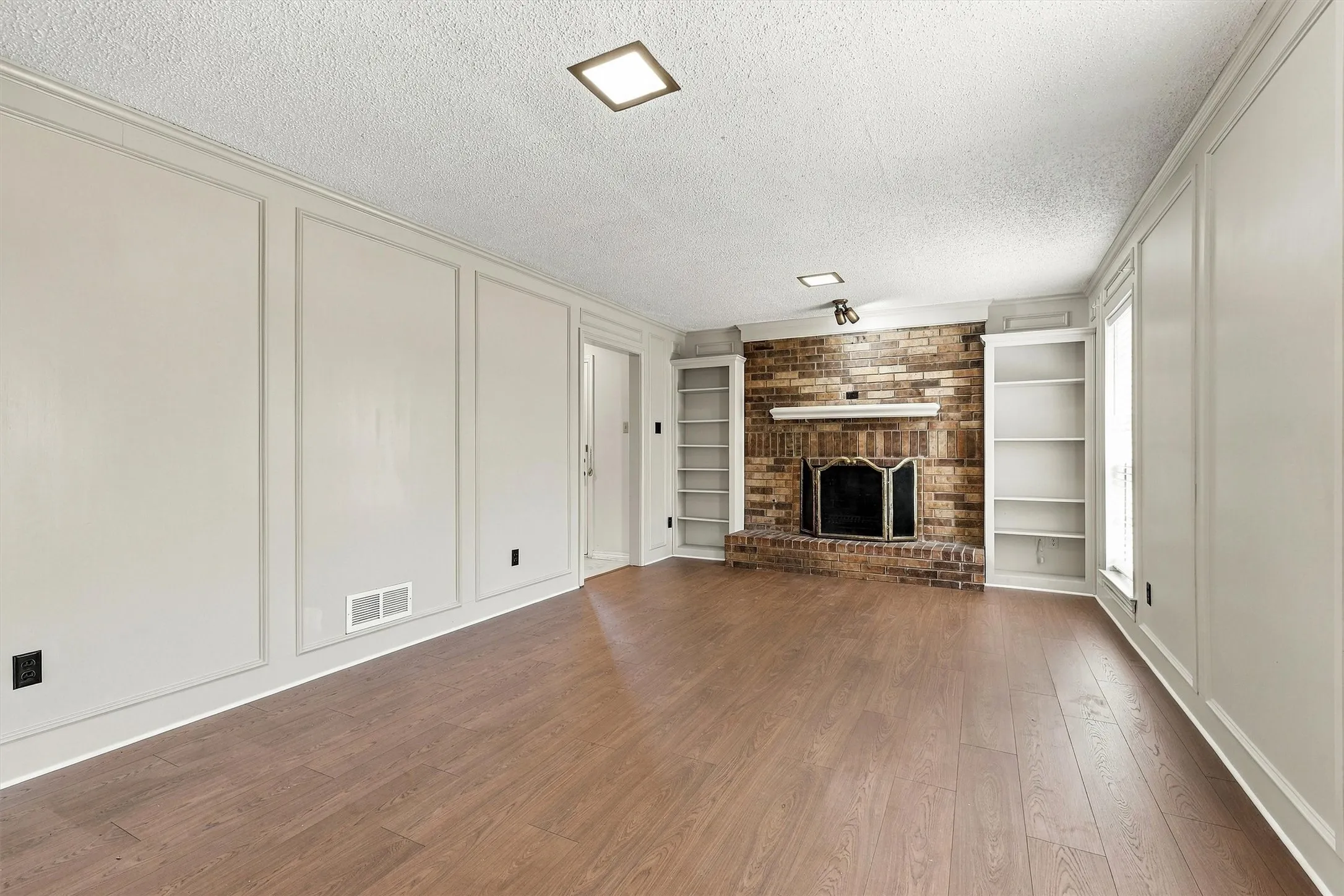 Living space featuring wood-finish flooring, floor-to-ceiling wainscoting, brick fireplace with a mantle, and built-in shelving