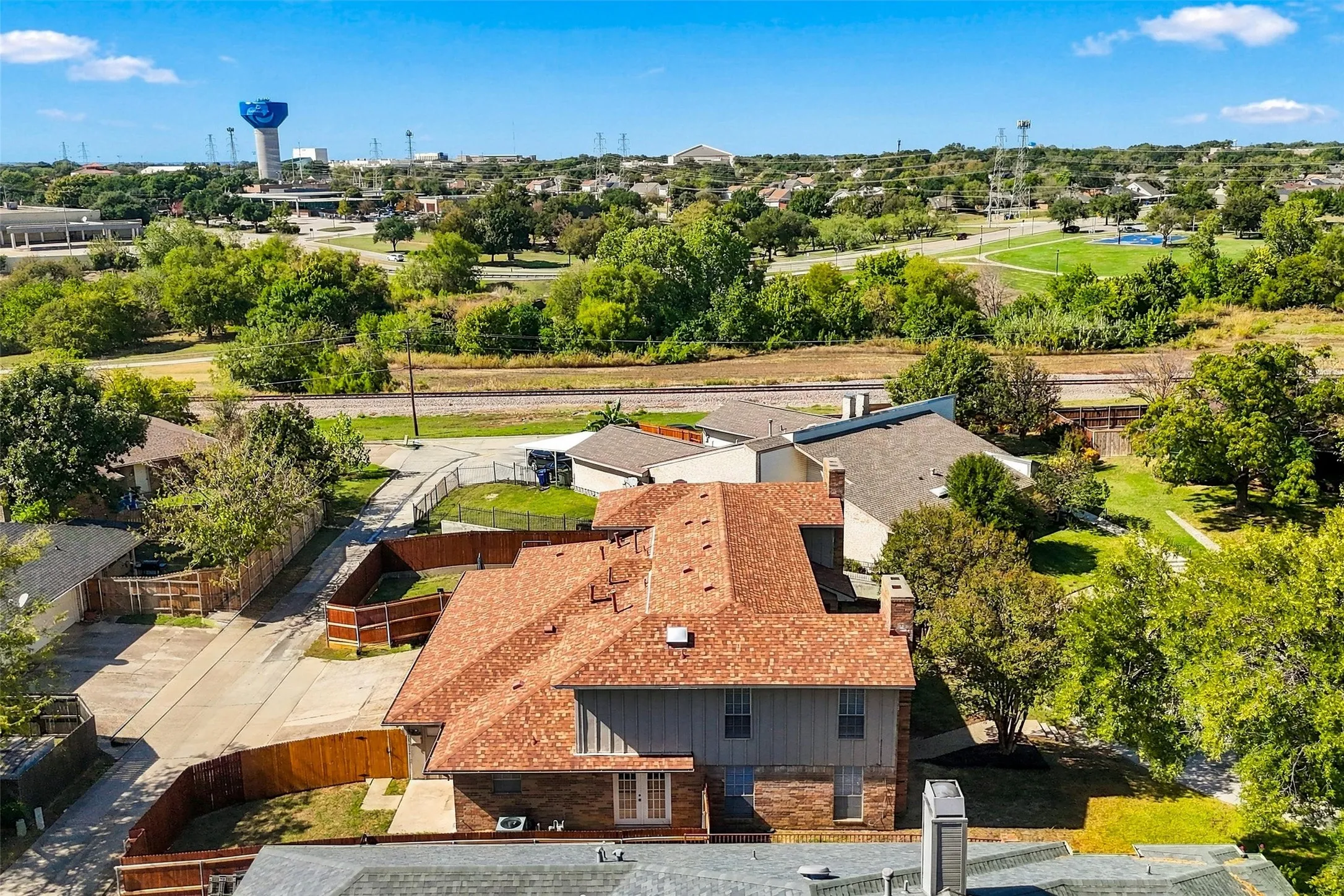 Aerial perspective of a multi-level structure featuring a multi-pitched composition shingle roof, brick and vertical siding exterior, wood-fenced perimeter, and dual masonry chimneys