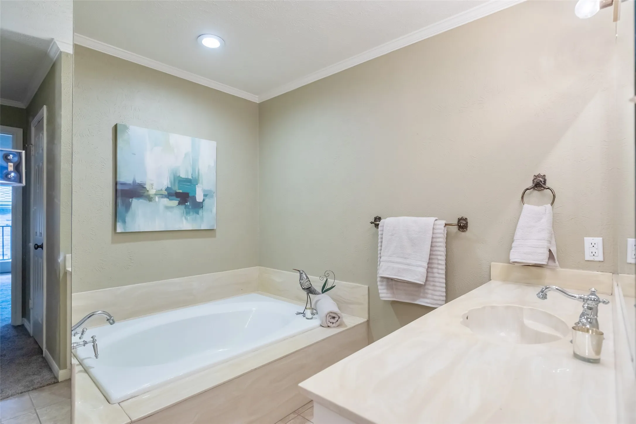 Built-in soaking tub with a tiled surround, vanity featuring an integrated basin, countertop with stone-pattern finish, and wall-mounted light fixtures