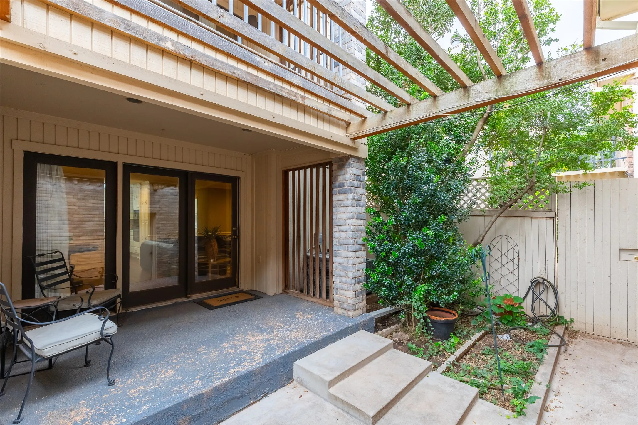 Covered patio featuring a wooden pergola, stone-accented pillar, glass sliding doors, and concrete steps leading to an enclosed outdoor area