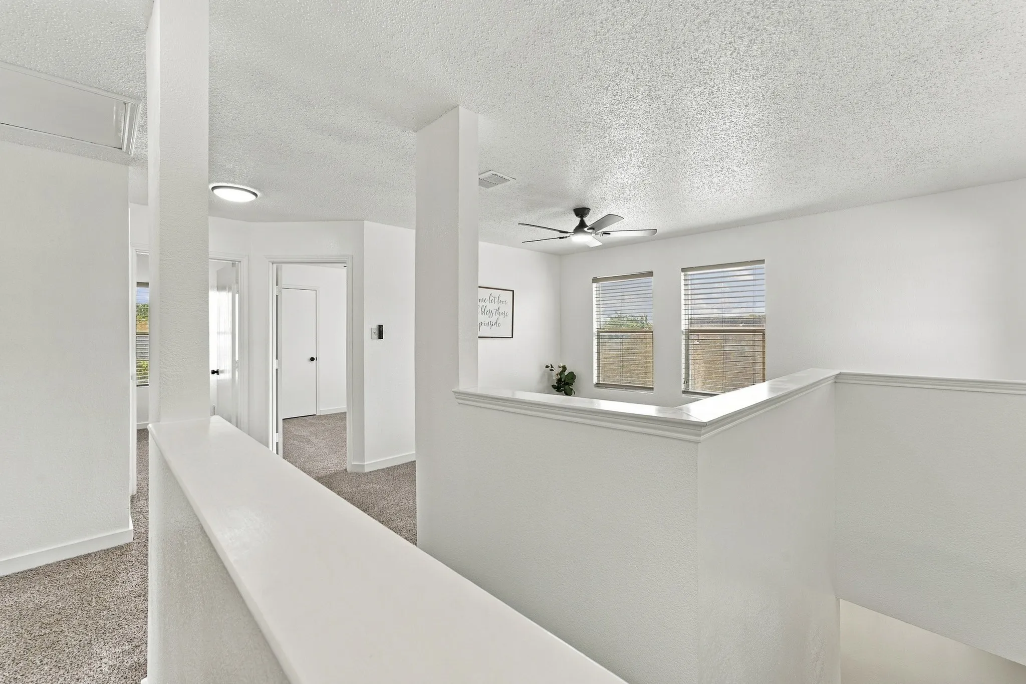Open floor plan interior featuring textured ceilings, carpeted flooring, a multi-blade ceiling fan with integrated lighting, and dual windows with horizontal blinds