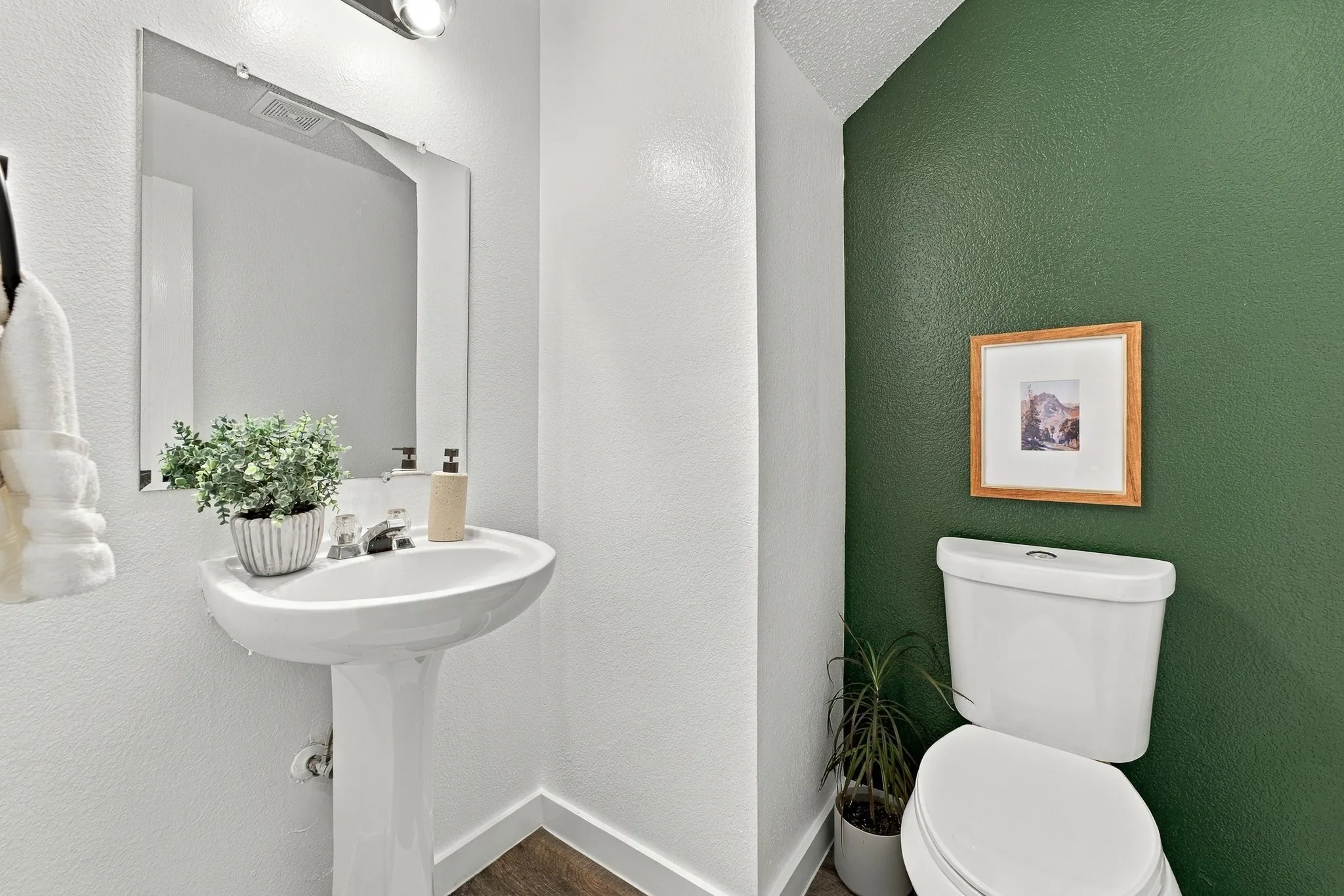Pedestal sink featuring a chrome faucet, frameless wall-mounted mirror, accent wall, and wood-finish flooring