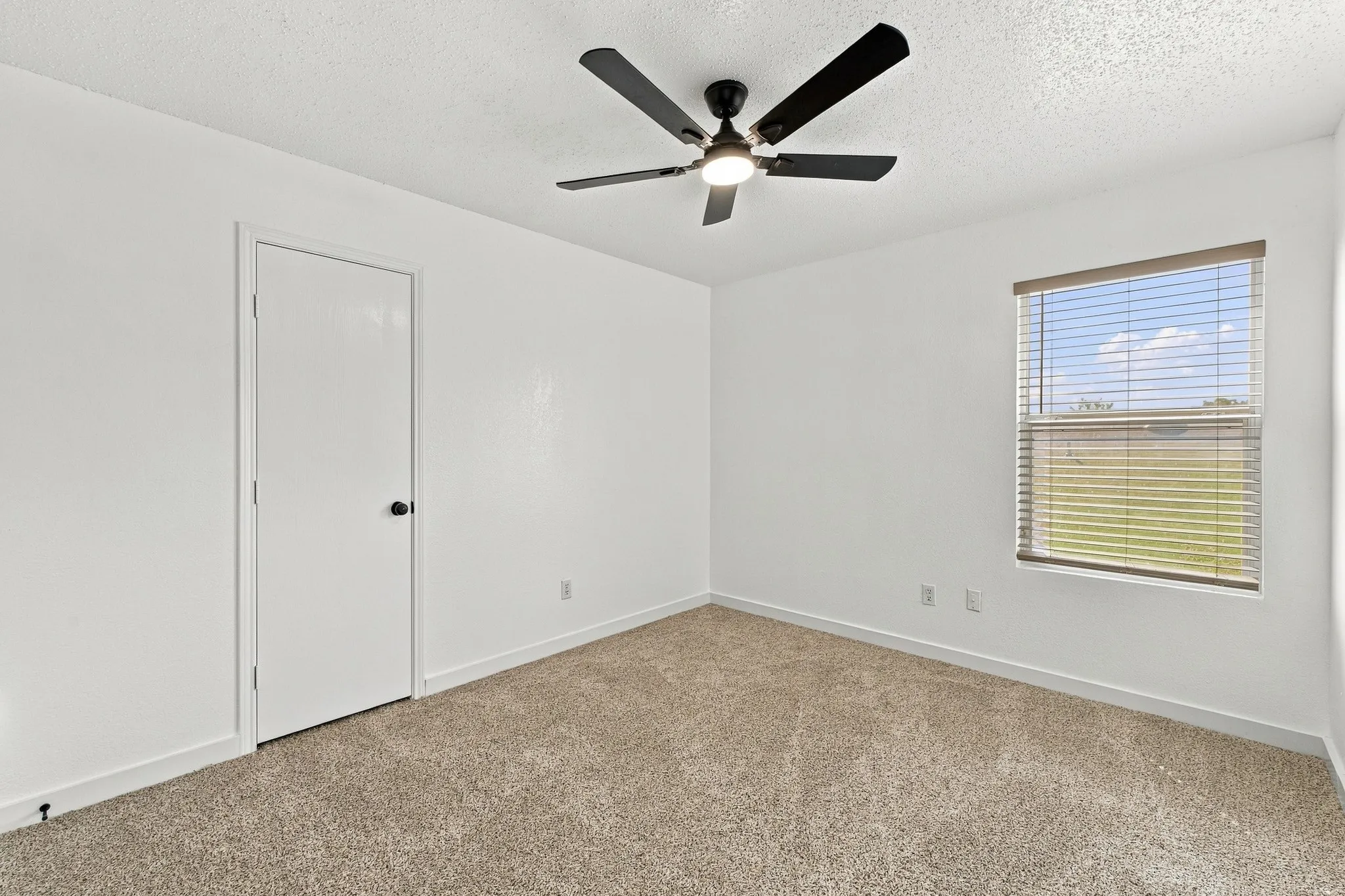 Neutral-toned carpeting, matte black ceiling fan with integrated lighting, single-panel interior door, and window with horizontal blinds