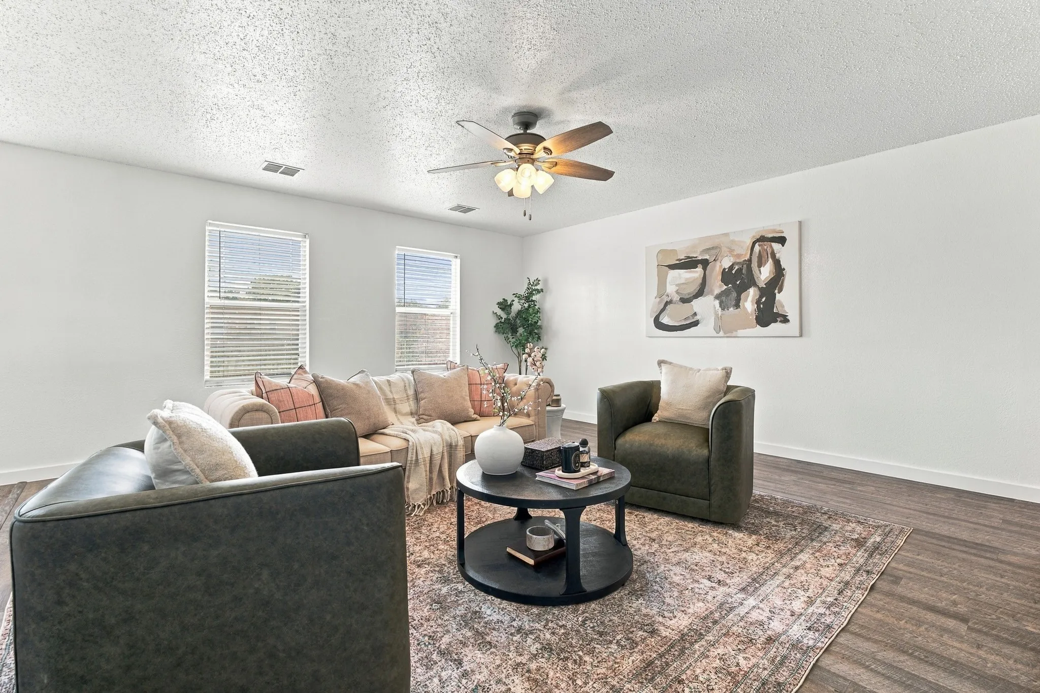 Living area featuring wood-finish flooring, two dual-pane windows with horizontal blinds, and a central ceiling fan with integrated lighting