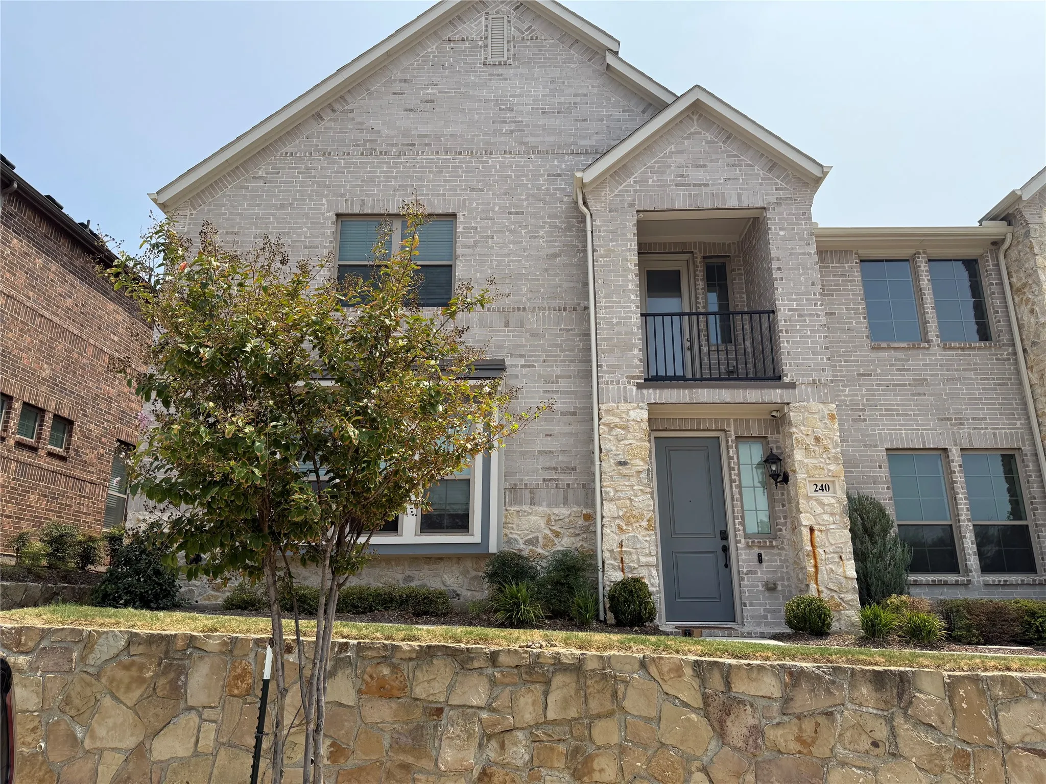 Two-story exterior featuring light-toned brick and stone veneer, a covered balcony with metal railing, and a flagstone retaining wall