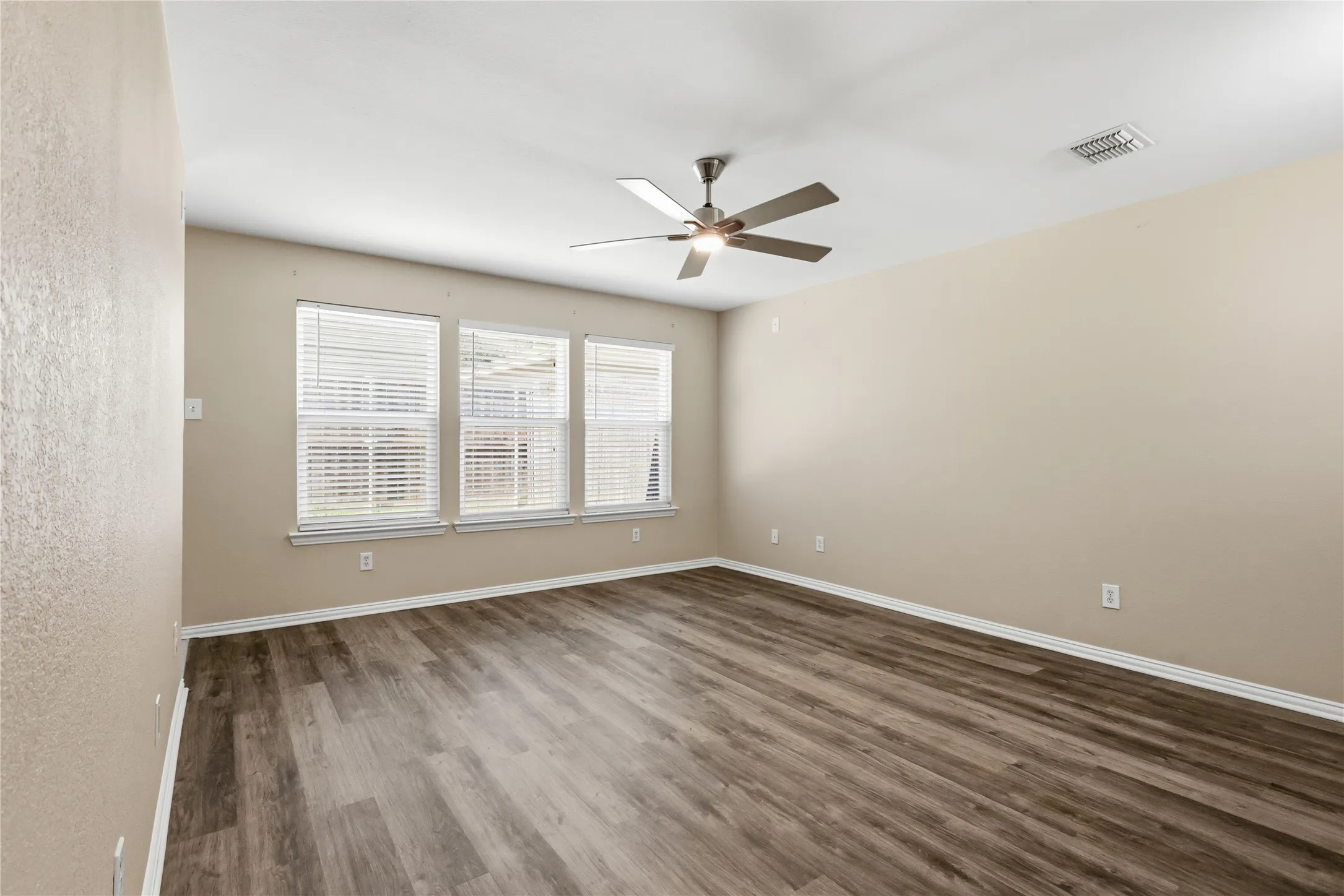 Neutral-toned walls featuring wood-finish flooring, a multi-blade ceiling fan, and three large windows with integrated blinds