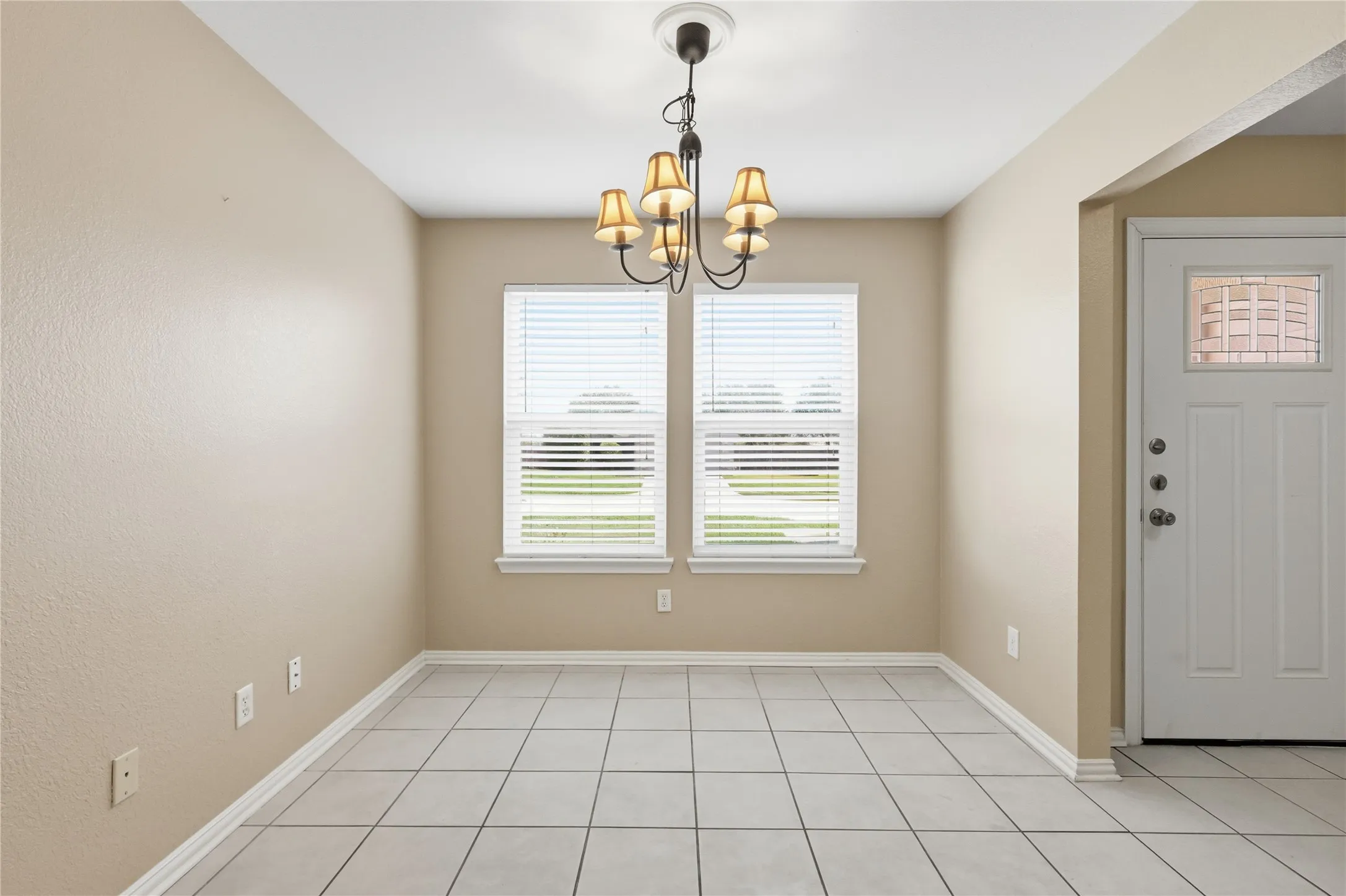 Ceramic tile flooring, multi-arm chandelier with fabric shades, dual windows with horizontal blinds, and a paneled entry door with a rectangular glass insert