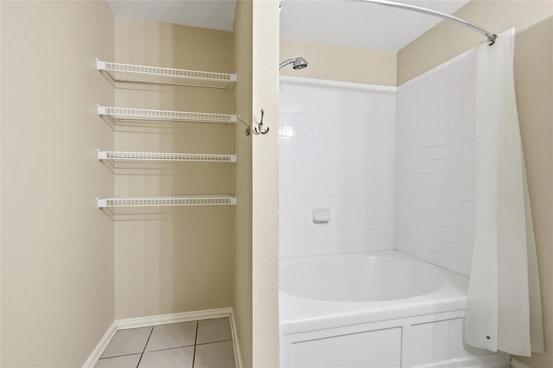 Bathroom featuring a built-in soaking tub with a white tile surround, a chrome shower head, and four wall-mounted storage shelves with tiled flooring
