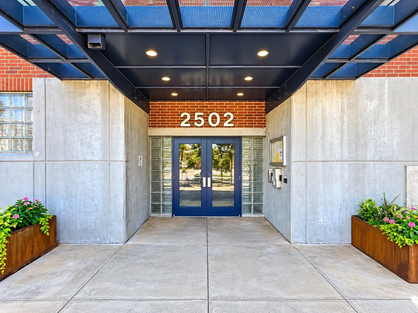 Industrial-style entryway featuring exposed brick masonry, smooth concrete panels, a structural metal portico, glass block sidelights, and double doors with blue metal frames