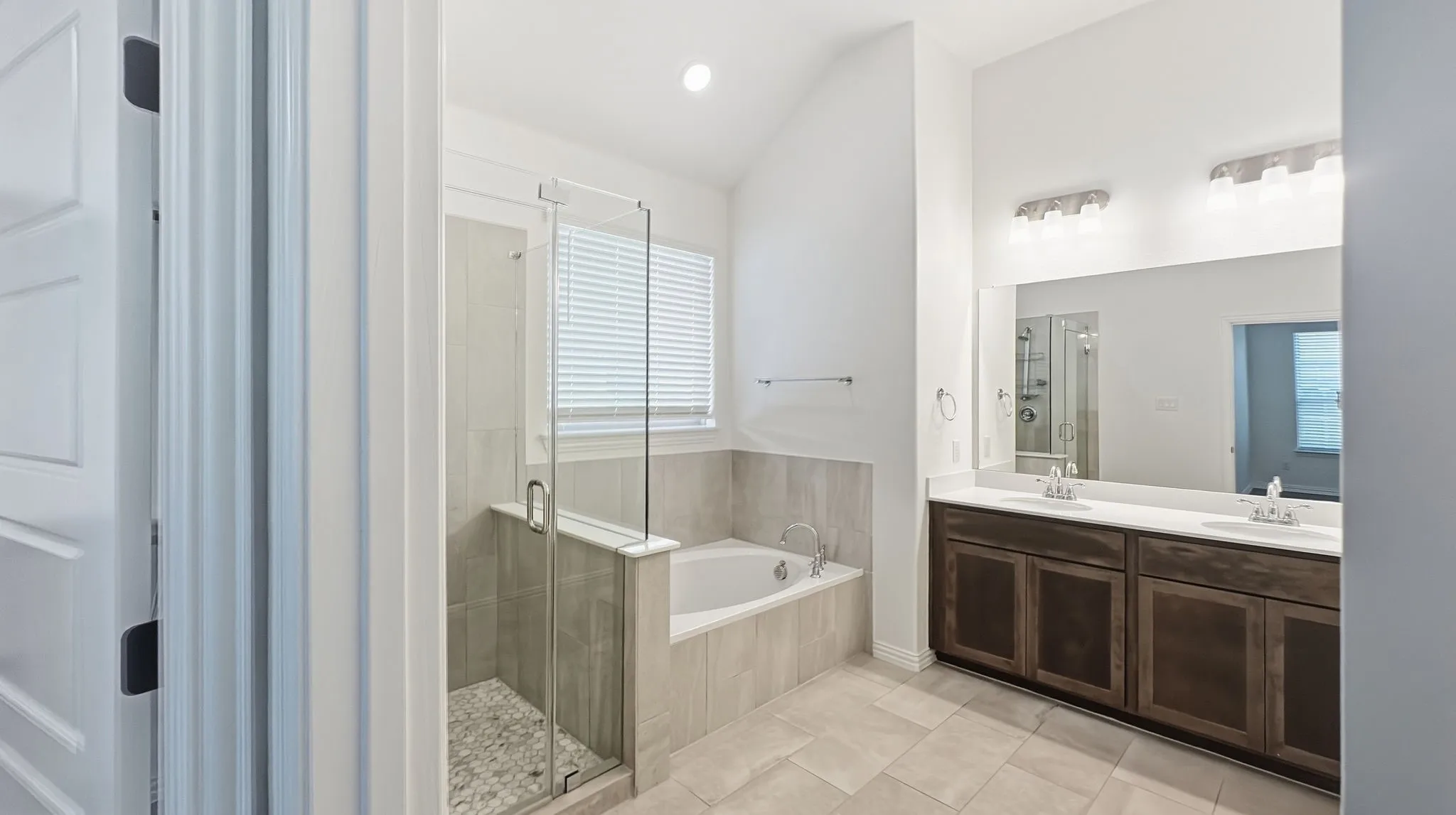 Bathroom featuring a dual-sink vanity with dark-finish cabinetry, a large frameless mirror, a deep soaking tub, a tiled walk-in shower with a glass enclosure, and high vaulted ceilings