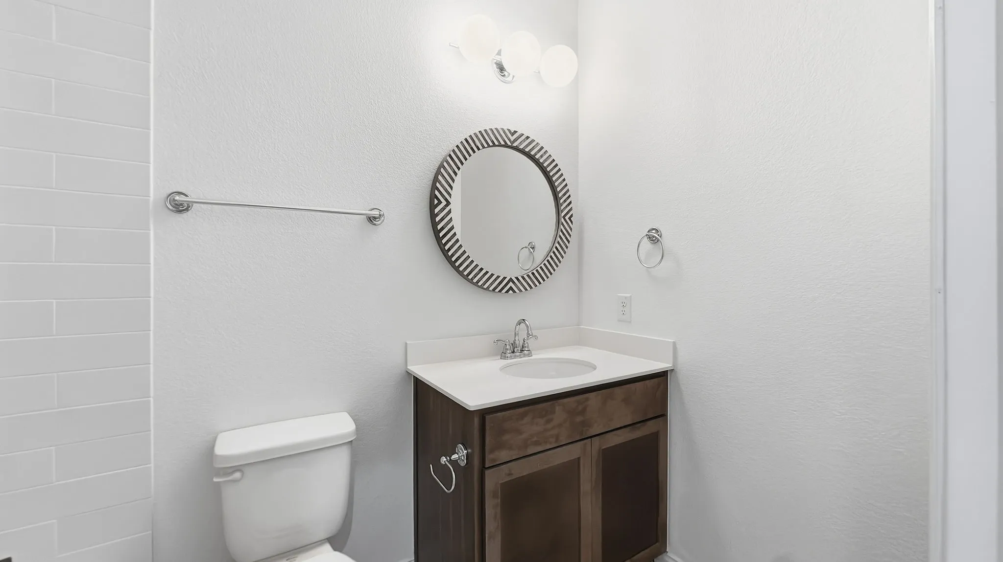 Vanity featuring wood-finish cabinetry, a white countertop with an integrated basin, a three-light globe vanity fixture, and a circular decorative wall mirror