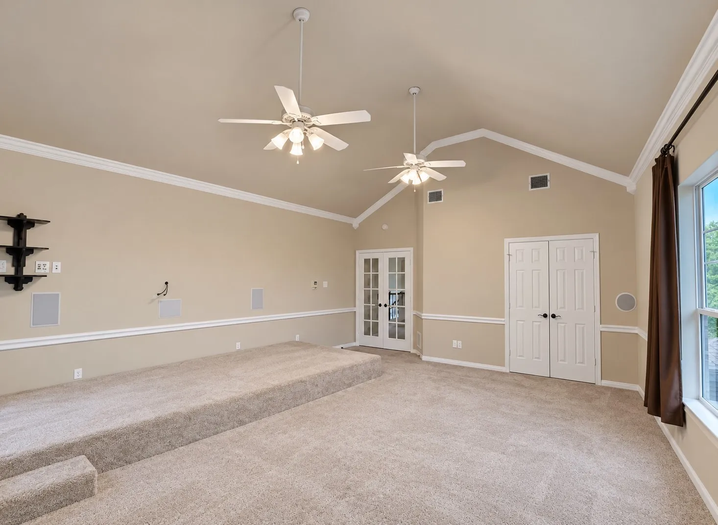 Vaulted ceiling with crown molding, two ceiling fans, built-in wall speakers, and a carpeted stepped platform