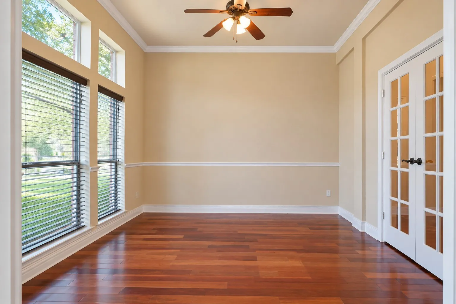 Rich wood-finish flooring, full-light French doors, crown molding, chair rail detailing, and a ceiling fan with integrated lighting