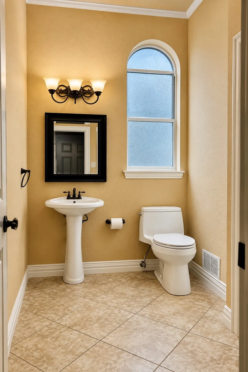 Pedestal sink, arched frosted window, tiled flooring, wall-mounted light fixture, crown molding