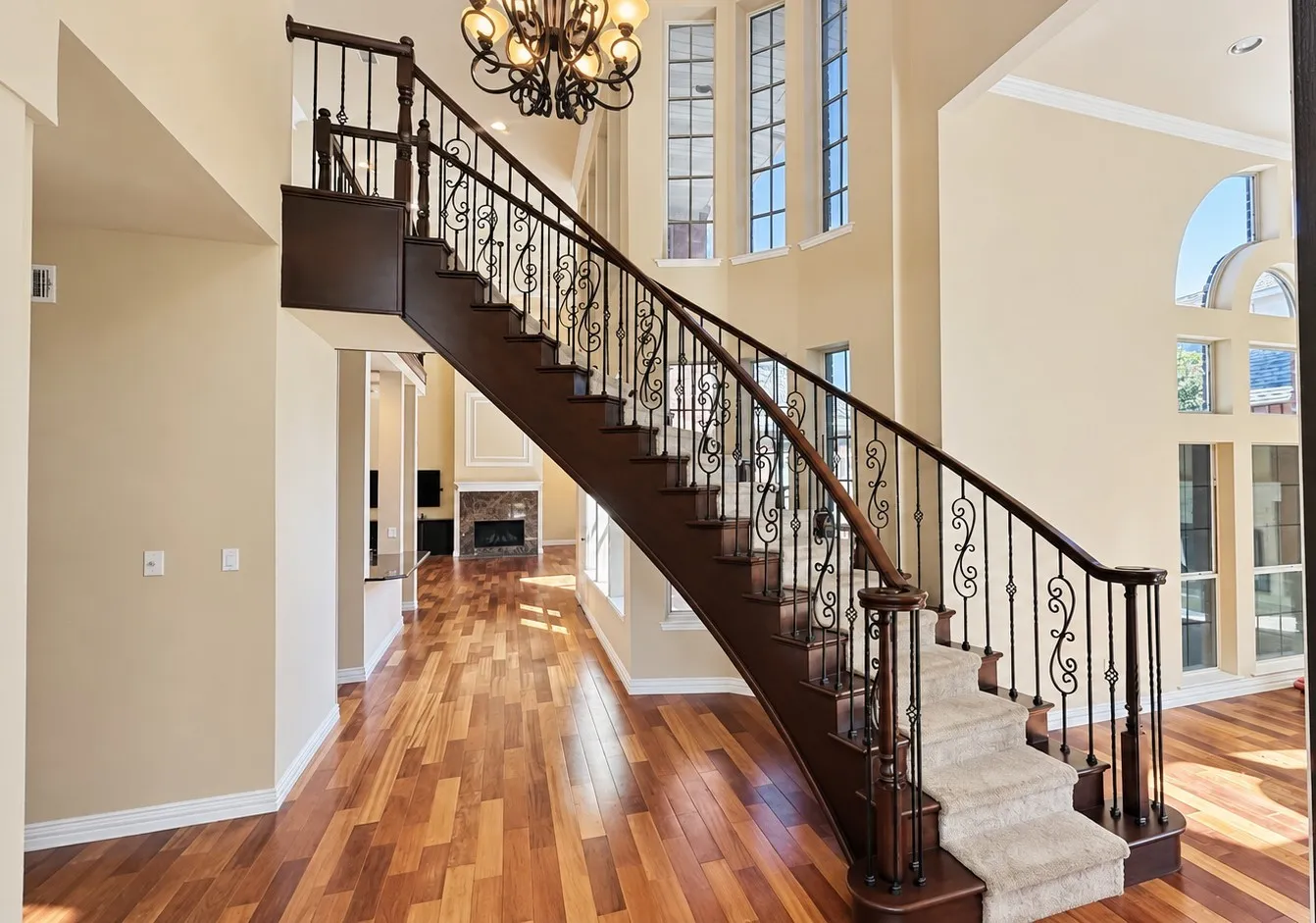Hardwood flooring, grand staircase with wrought iron balusters and carpeted treads, chandelier lighting fixture, and high ceilings with multiple windows