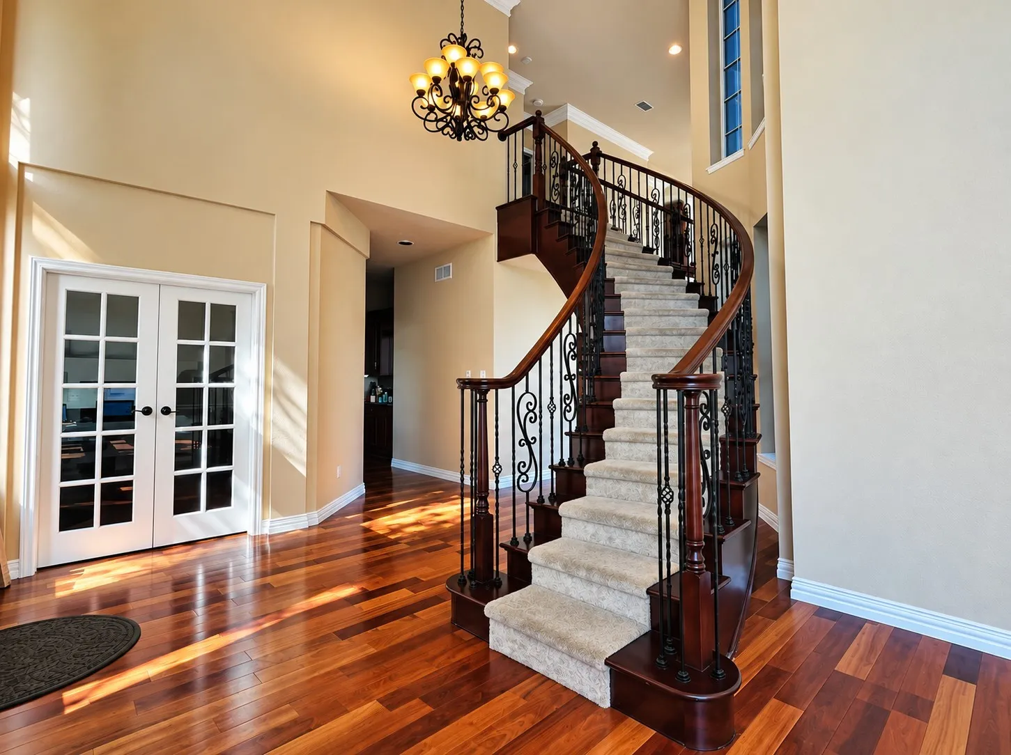 Grand curved staircase with wrought iron balusters and wood-finish handrail, wood-finish flooring, double French doors with grid panes, and a decorative chandelier