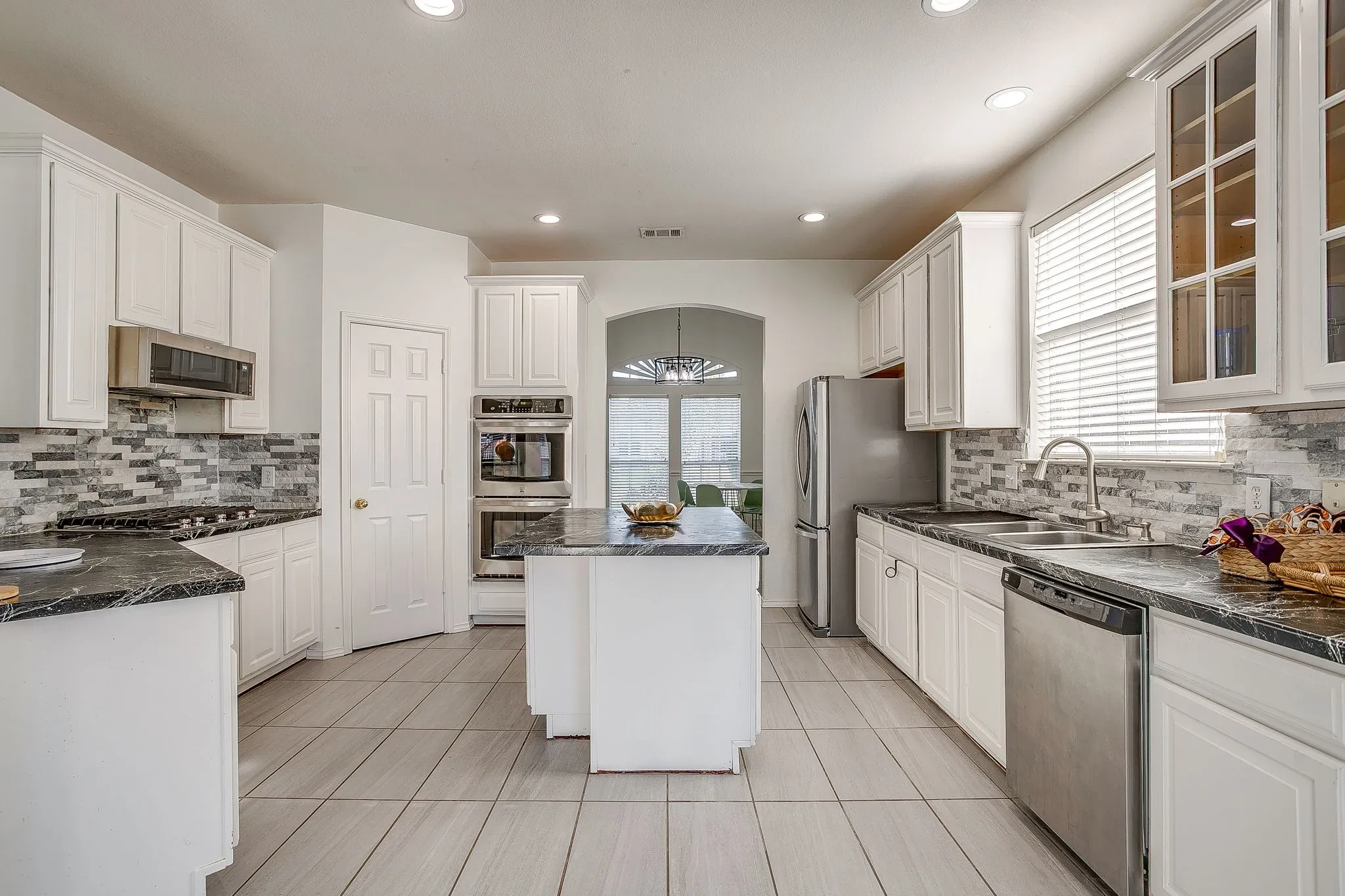 Kitchen featuring white cabinetry, a central island with a stone-finish countertop, a multi-textured tile backsplash, built-in stainless steel double ovens, and tile flooring