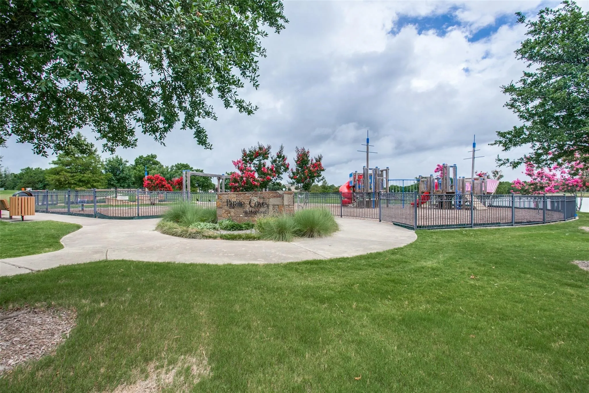 Community playground featuring structured play equipment, iron perimeter fencing, concrete pathways, and manicured green space