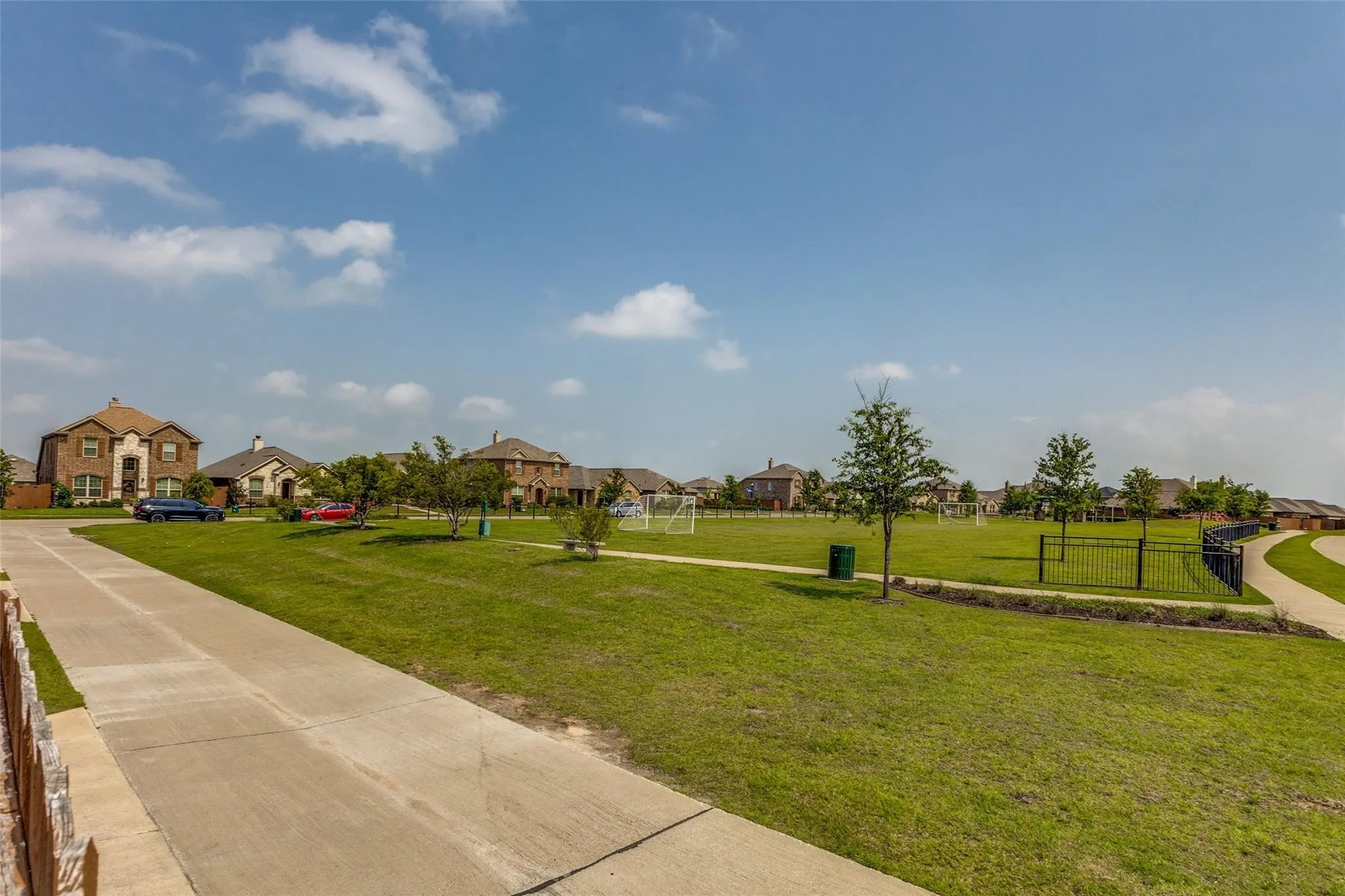 Expansive green space featuring a concrete pathway, mature trees, and metal perimeter fencing