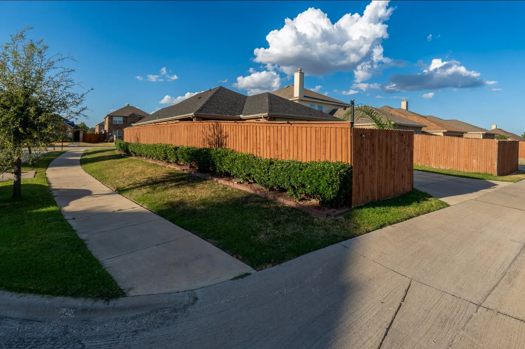 Corner lot exterior featuring a wood privacy fence, concrete sidewalk, manicured hedging, and grass lawn