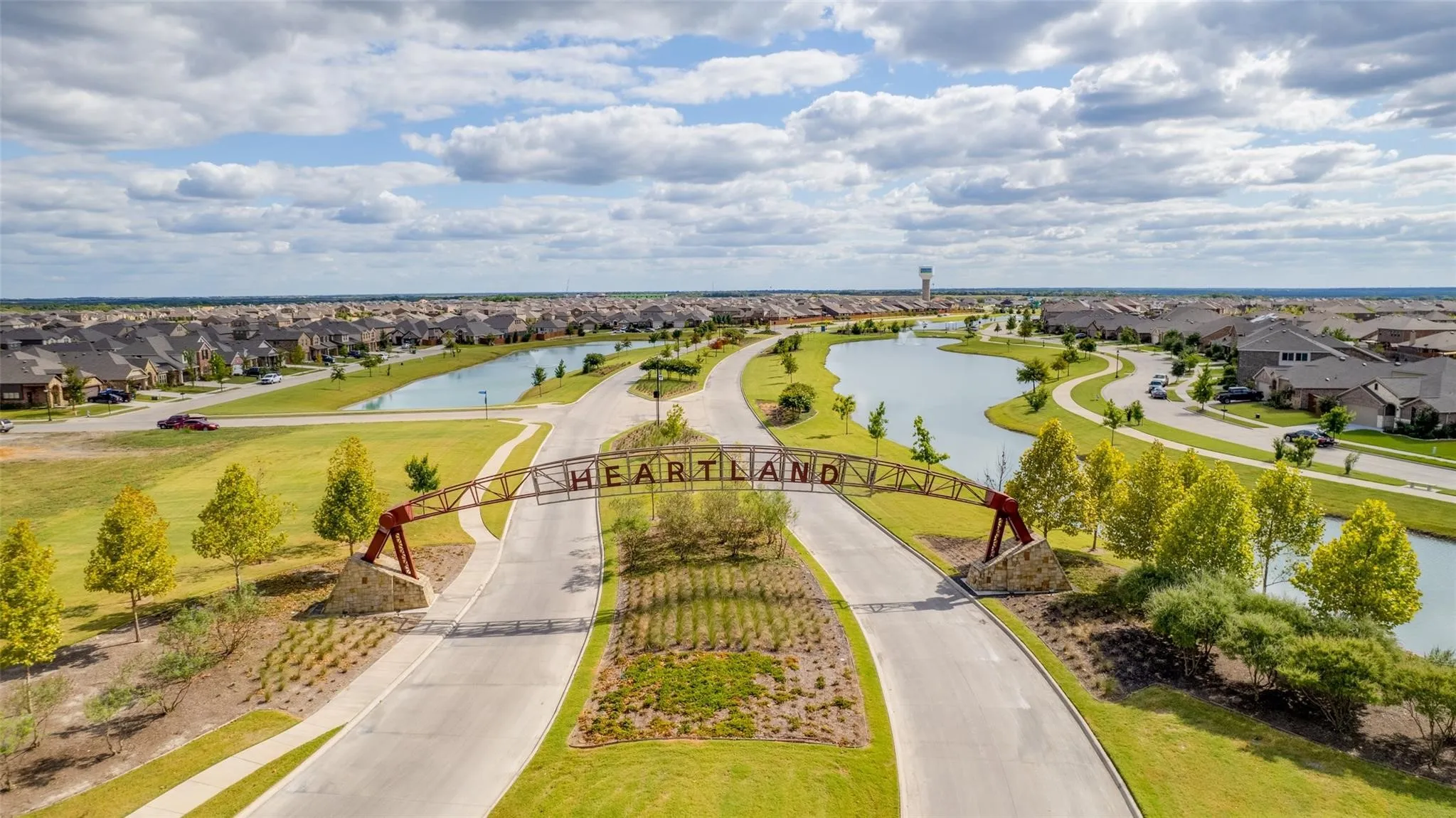 Community entrance featuring a metal architectural archway, stone pillar foundations, paved roadways, retention ponds, and landscaped green space
