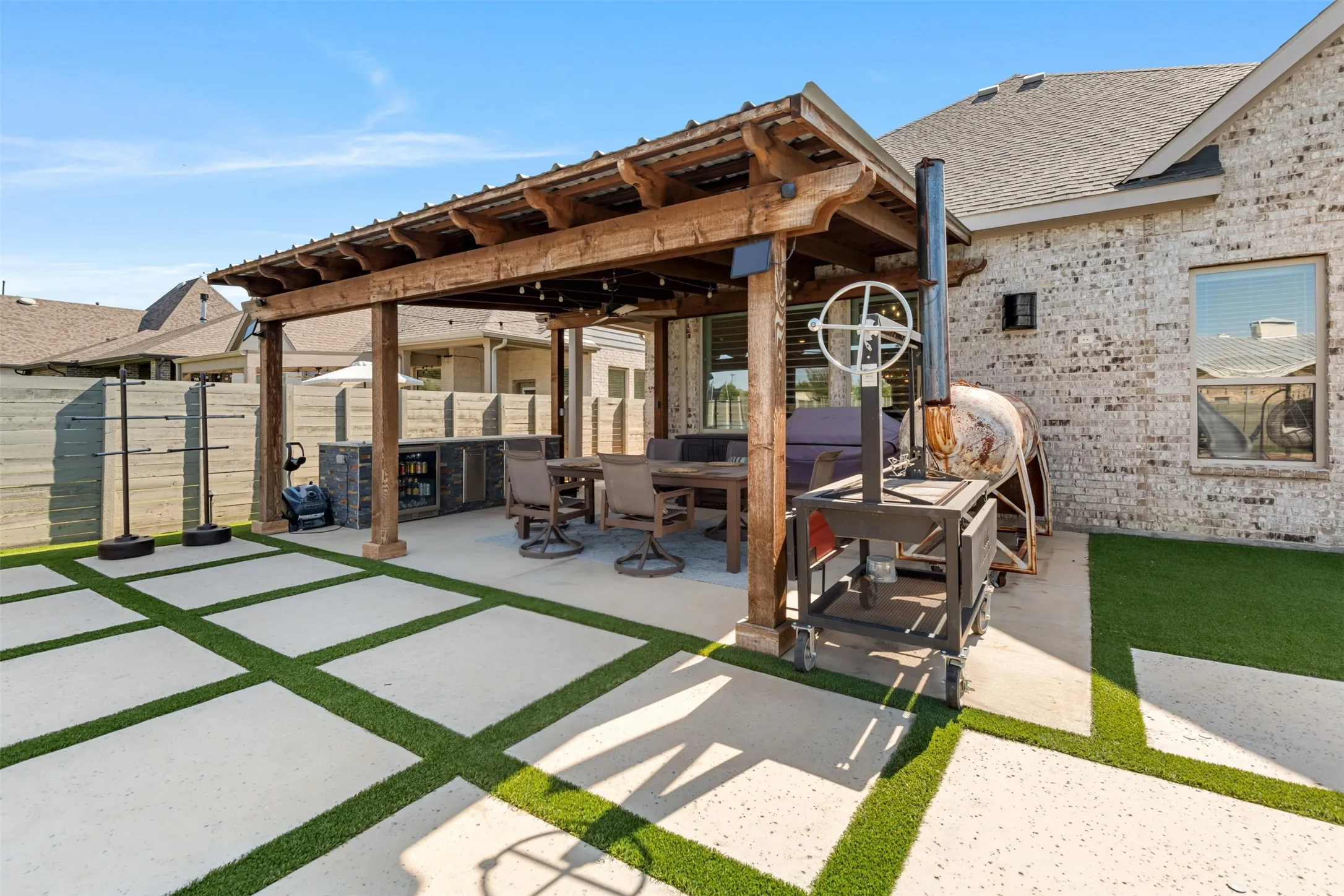 Covered patio featuring a timber-frame pergola, an integrated outdoor kitchen with stainless steel built-in cabinetry, and large-format concrete pavers with turf inlays