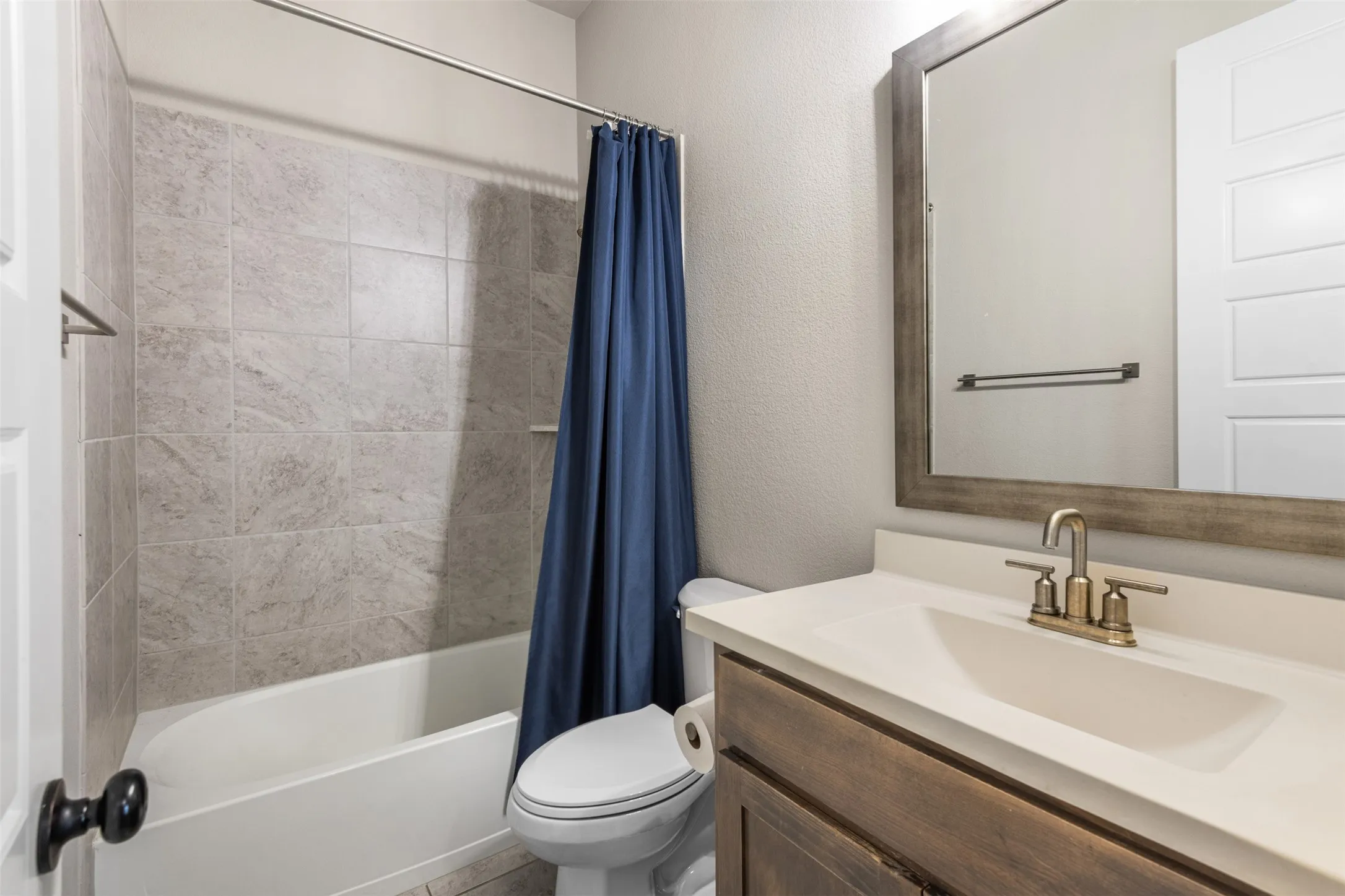 Bathroom featuring a wooden vanity cabinet, solid-surface countertop with integrated sink, brushed brass faucet, framed wall mirror, and a tub-shower combination with ceramic tile surround