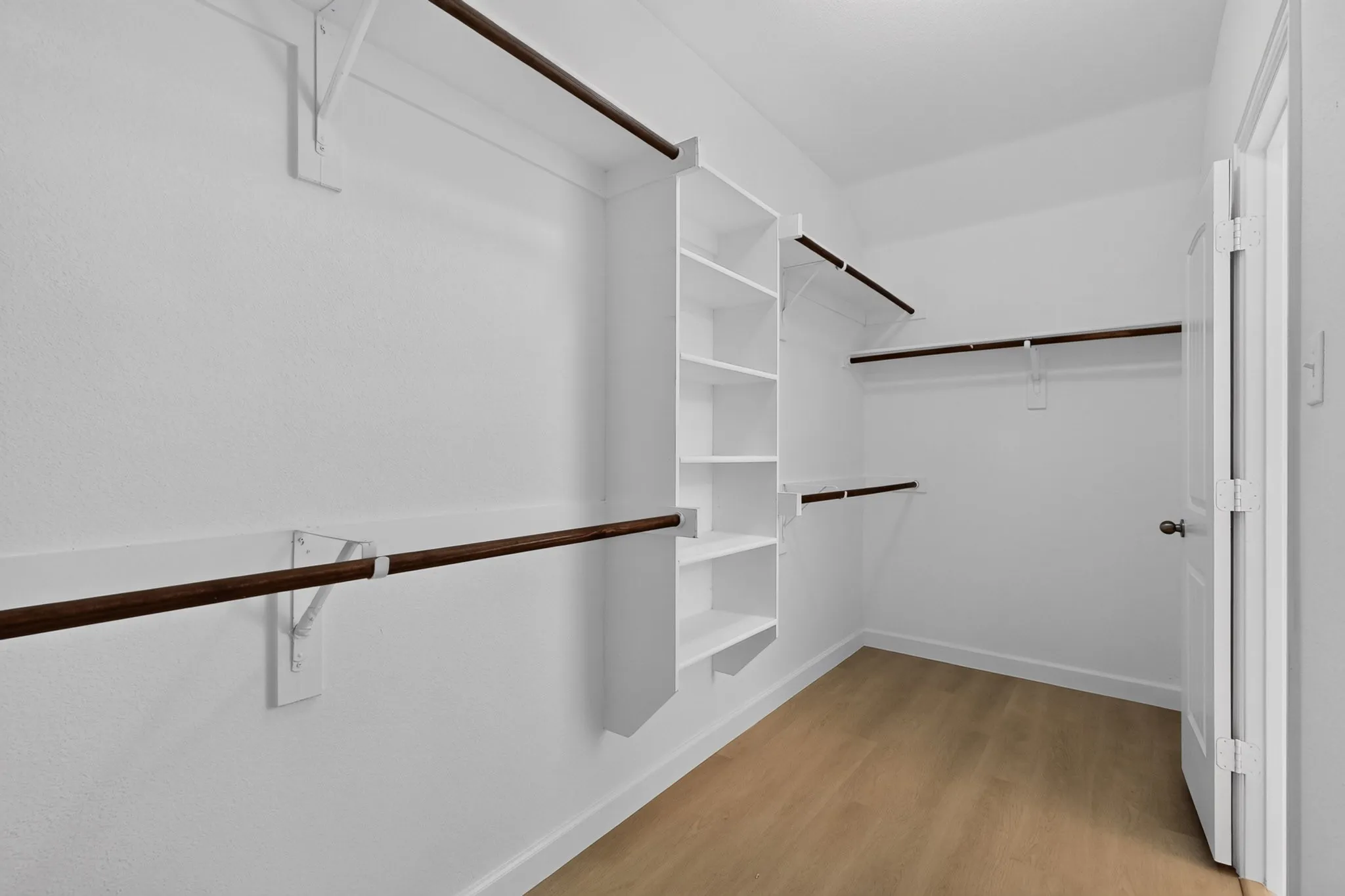 Walk-in storage space featuring wood-finish flooring, white built-in shelving, and dark-toned hanging rods